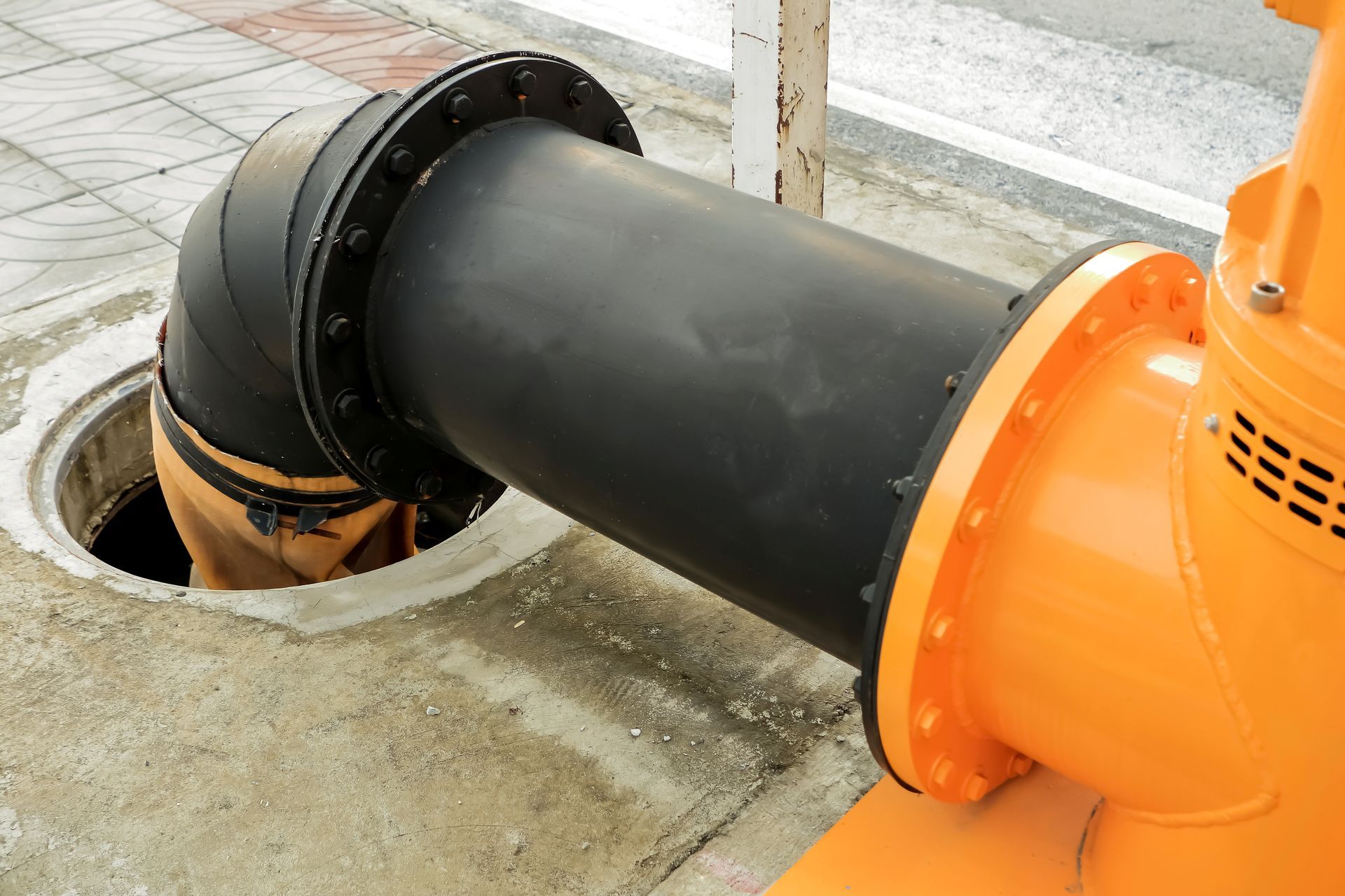 Black pipe connected to orange industrial machinery over a concrete drainage opening. Black pipe connected to orange industrial equipment beside a concrete drain opening