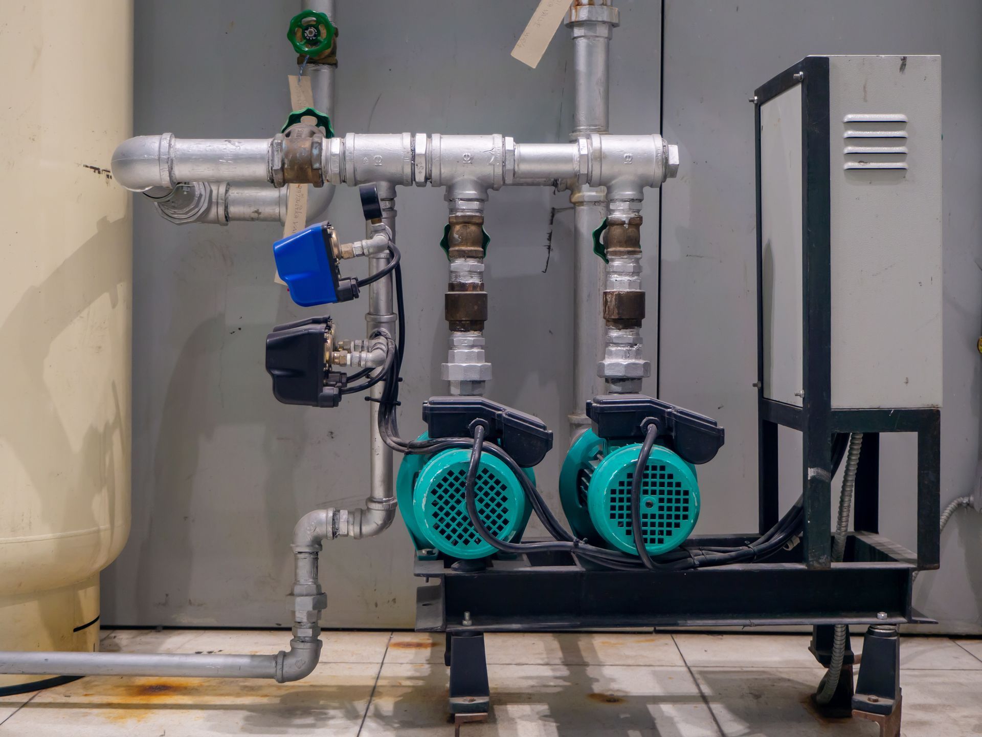 Industrial pump system with two turquoise motors, silver pipes, and a control box against a wall