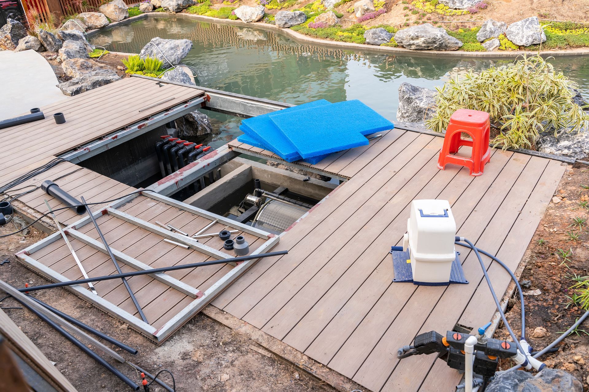 Backyard deck with an open hatch, pond beyond, and stacked chairs, a blue cushion, and a red stool