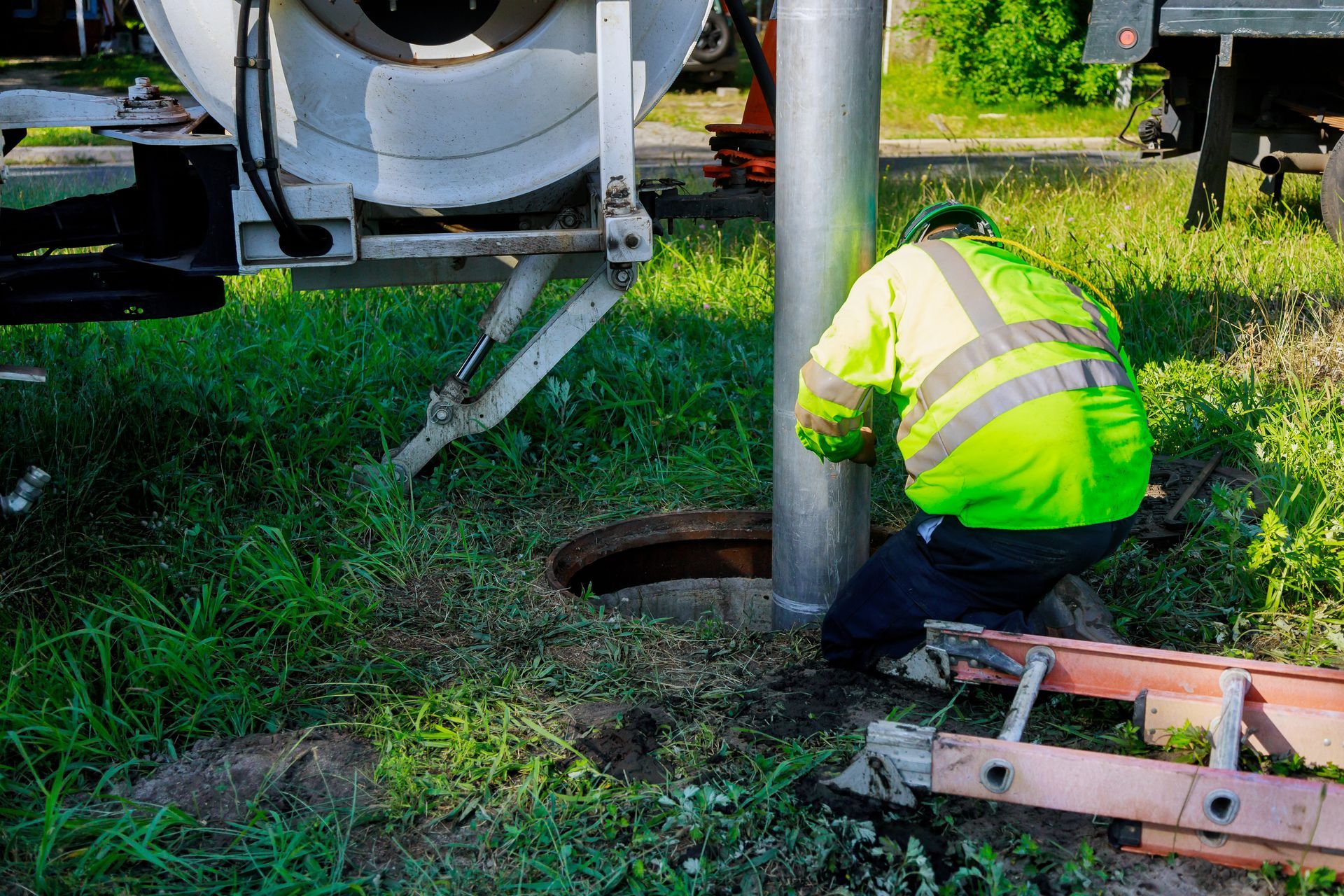 Worker in neon safety jacket kneels beside a concrete truck, inspecting a hole in grassy ground.