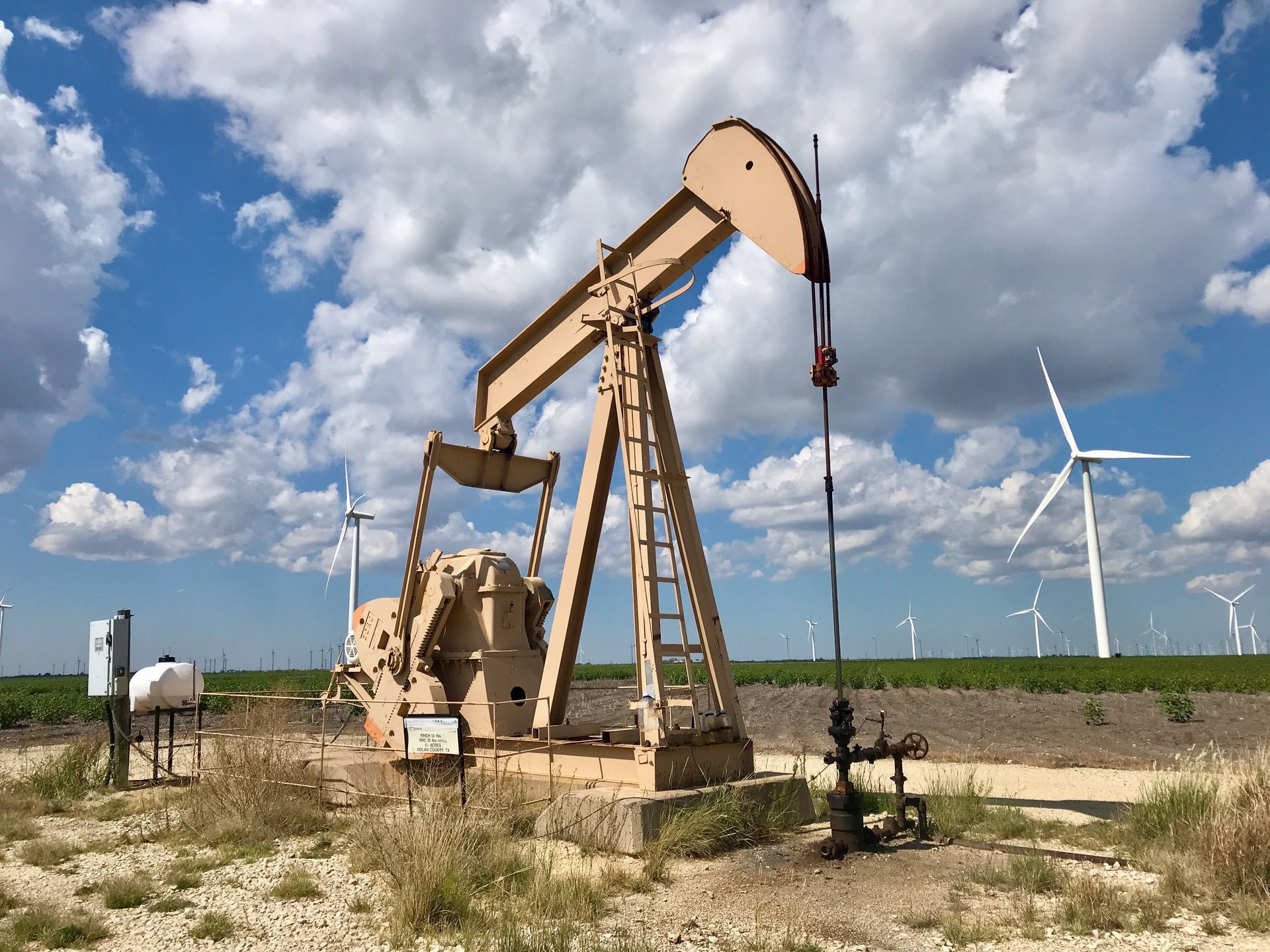 Pumpjack oil well in a grassy field with wind turbines under a cloudy blue sky