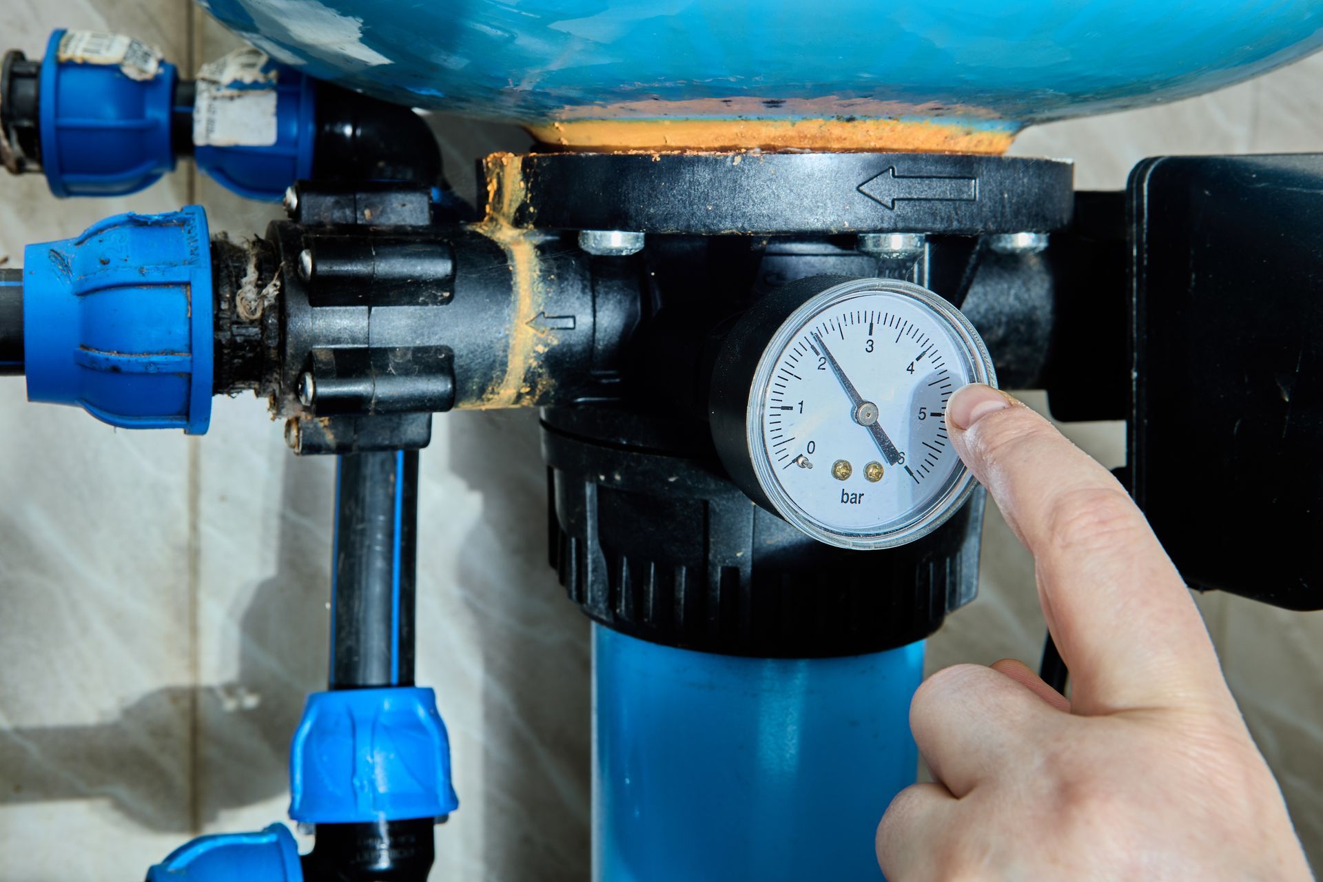 Hand adjusting a pressure gauge on blue plumbing pipes and a black filter unit