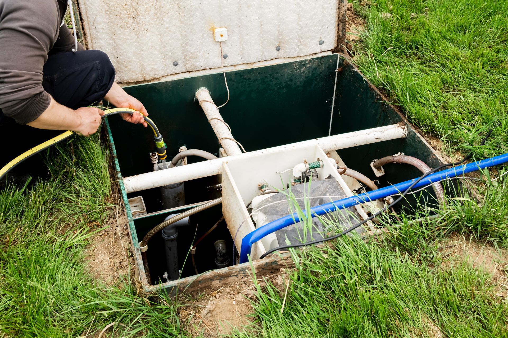 Person inspecting pipes in an open underground utility box beside grass.