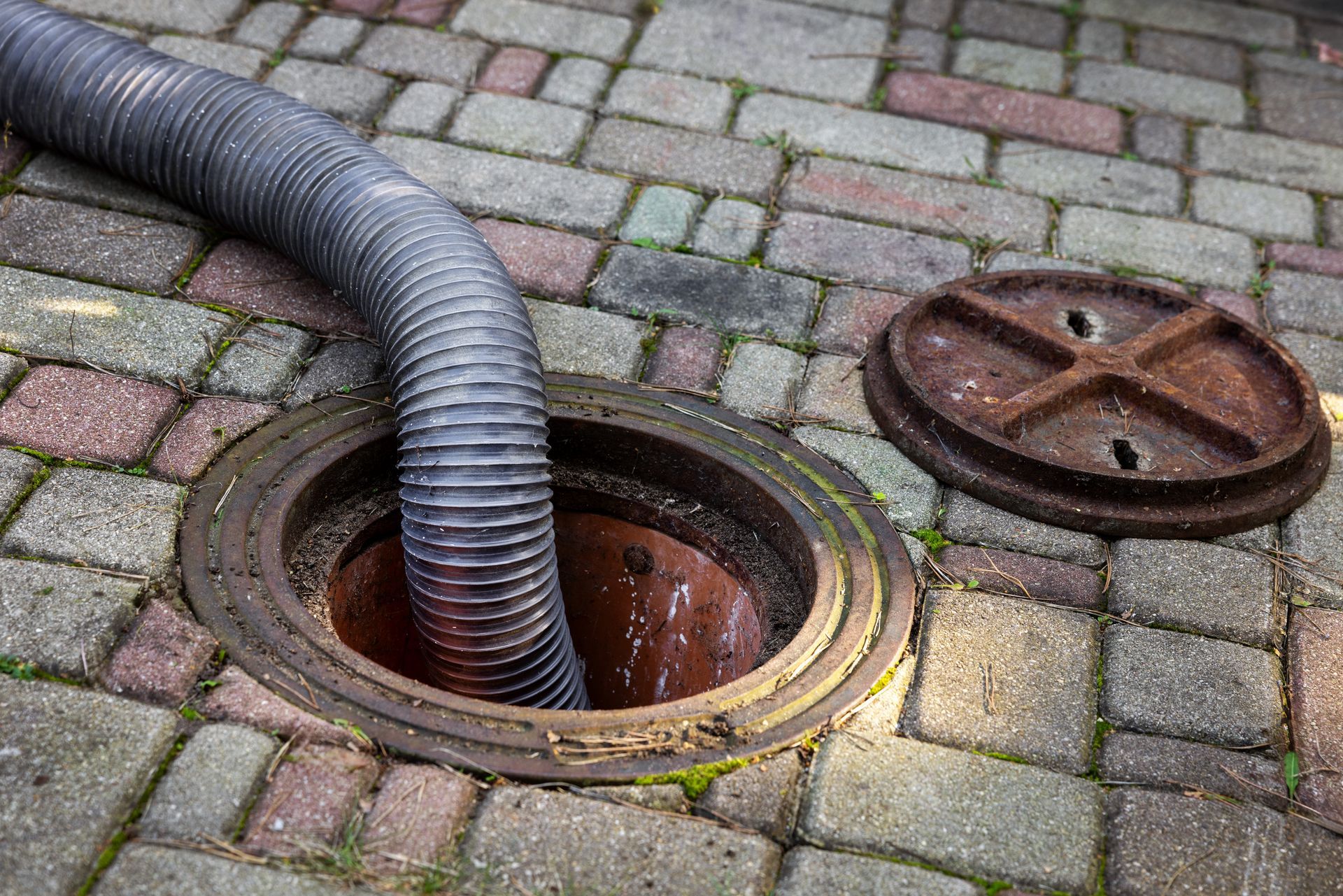 Gray hose inserted into an open manhole cover on a brick-paved street