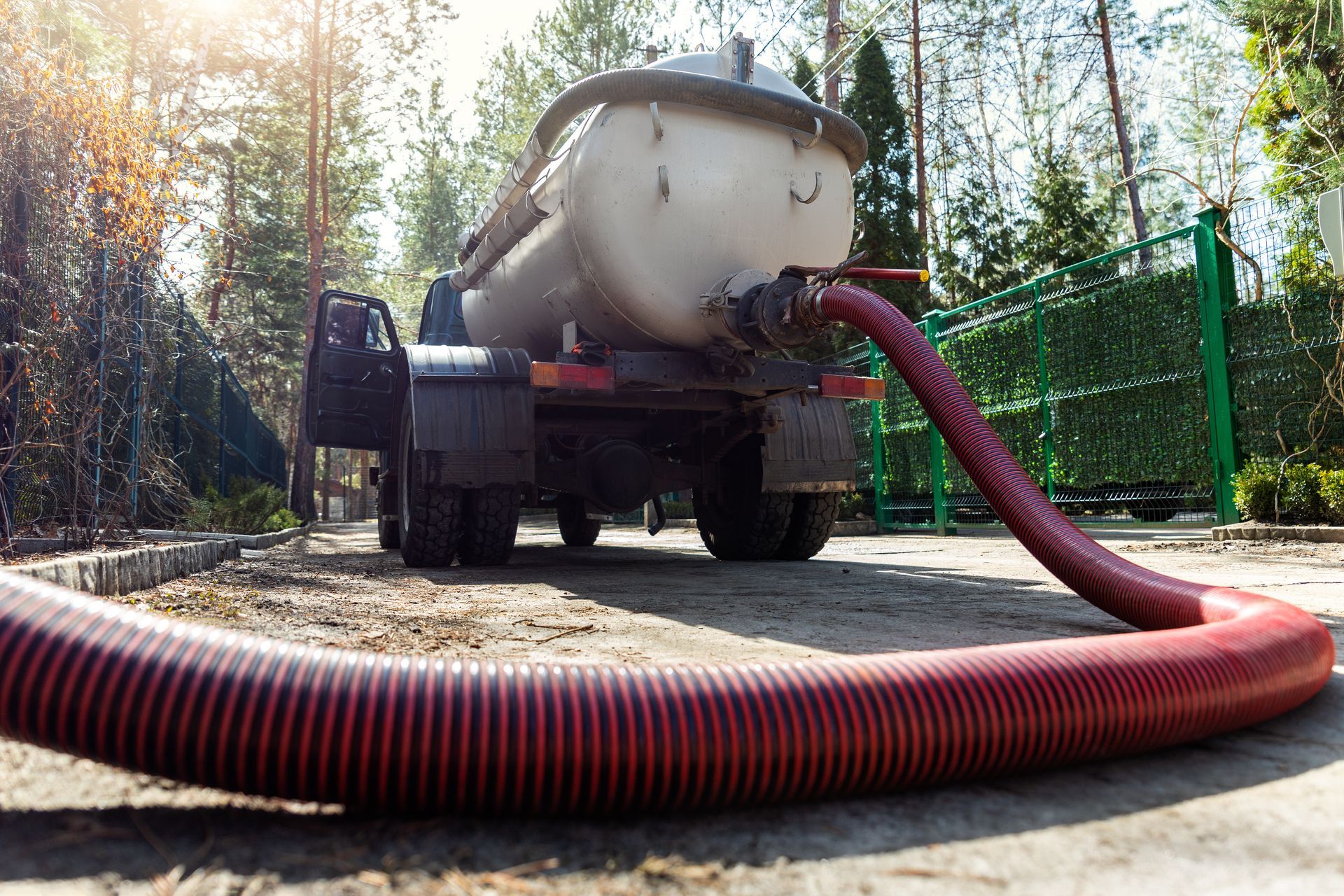 Truck-mounted vacuum hose on a rural road beside a large tank, with forest and fence in the background