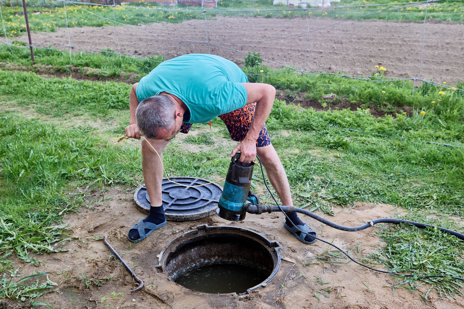 Person in turquoise shirt crouches by an open manhole, using a hose and pump outdoors.