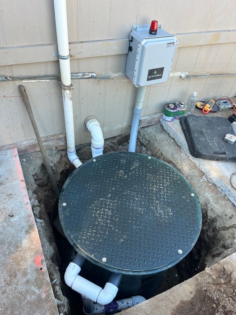 Open utility pit with black round cover, white pipes, and gray electrical box against a building wall.