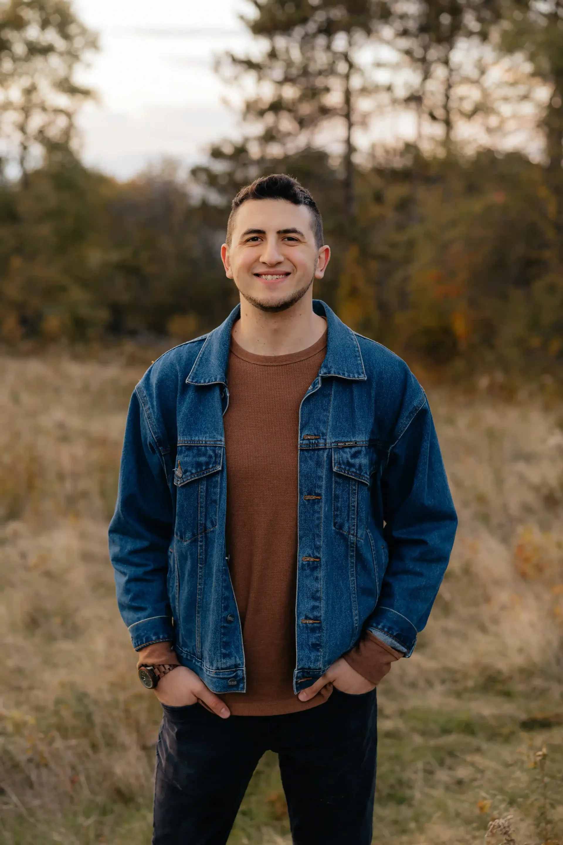 A man in a denim jacket and sweater is standing in a field with his hands in his pockets.