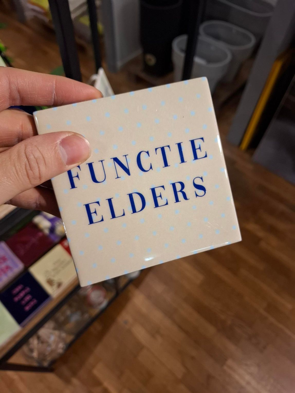 Hand die een beige kaartje met blauwe tekst vasthoudt waarop 'FUNCTIE ELDERS' staat, in een winkelgang.