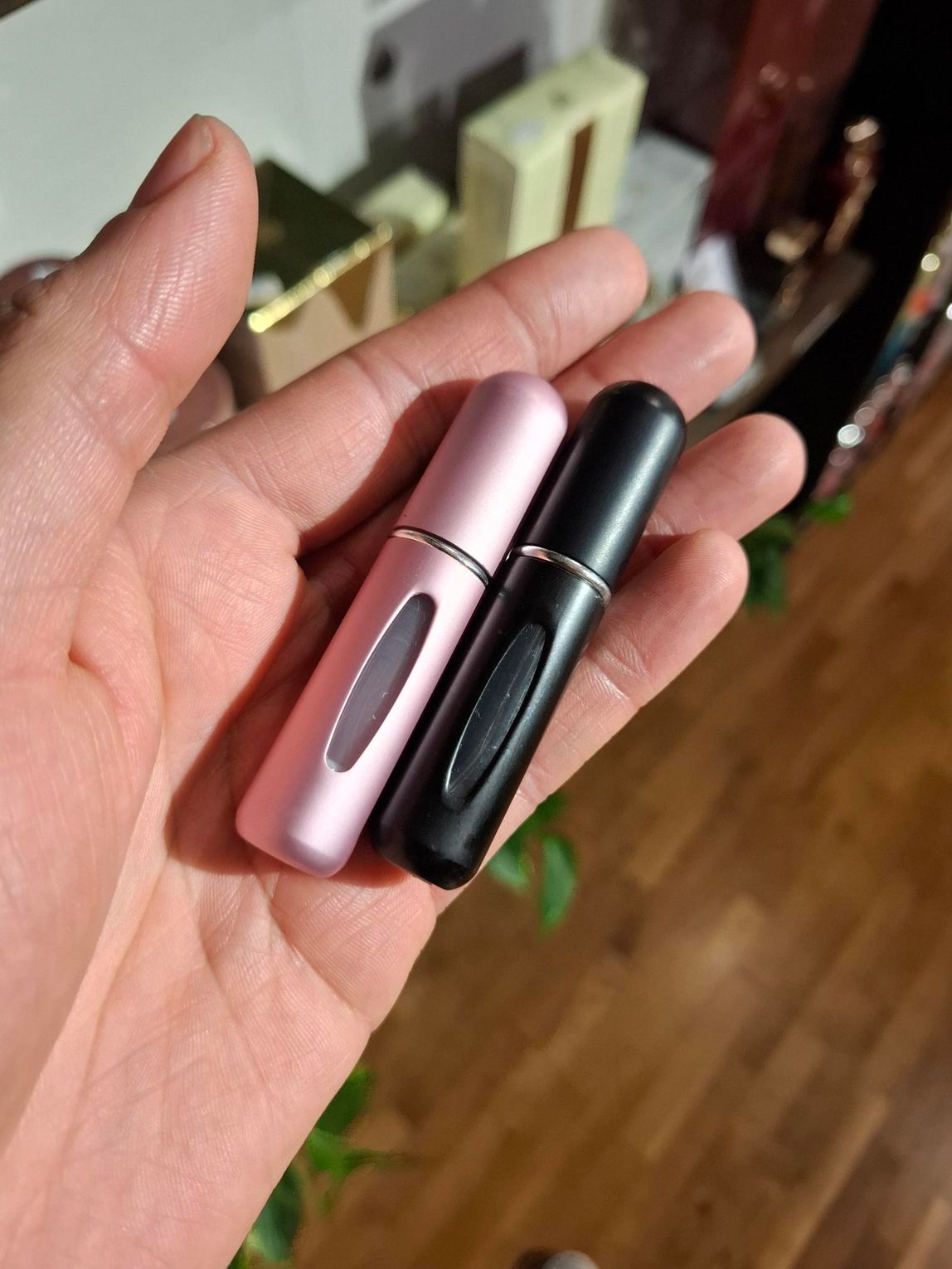 Een hand houdt twee kleine, cilindrische parfumverstuivers vast: een roze en een zwarte, beide met een verticaal kijkvenster.