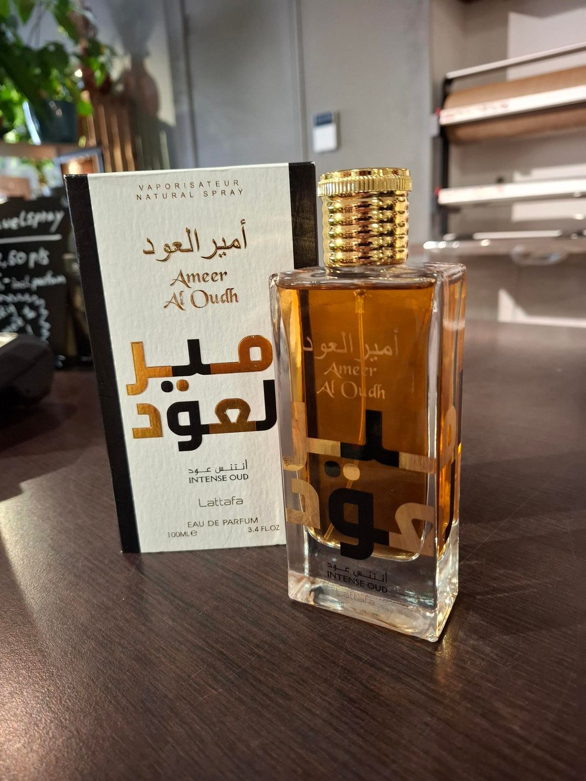 Een flesje Ameer Al Oudh-parfum staat naast de wit-gouden doos op een houten tafel.
