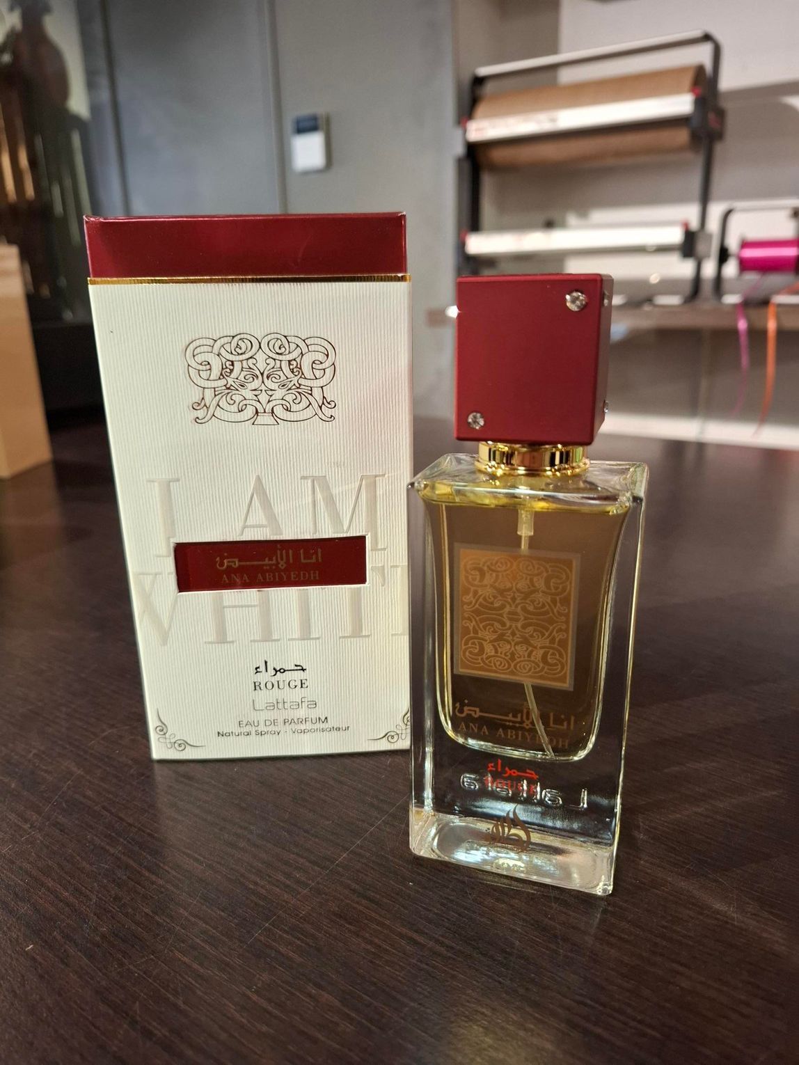 Een parfumflesje en de bijbehorende rechthoekige doos met witte en donkerrode merkopdruk op een donkerhouten tafel.