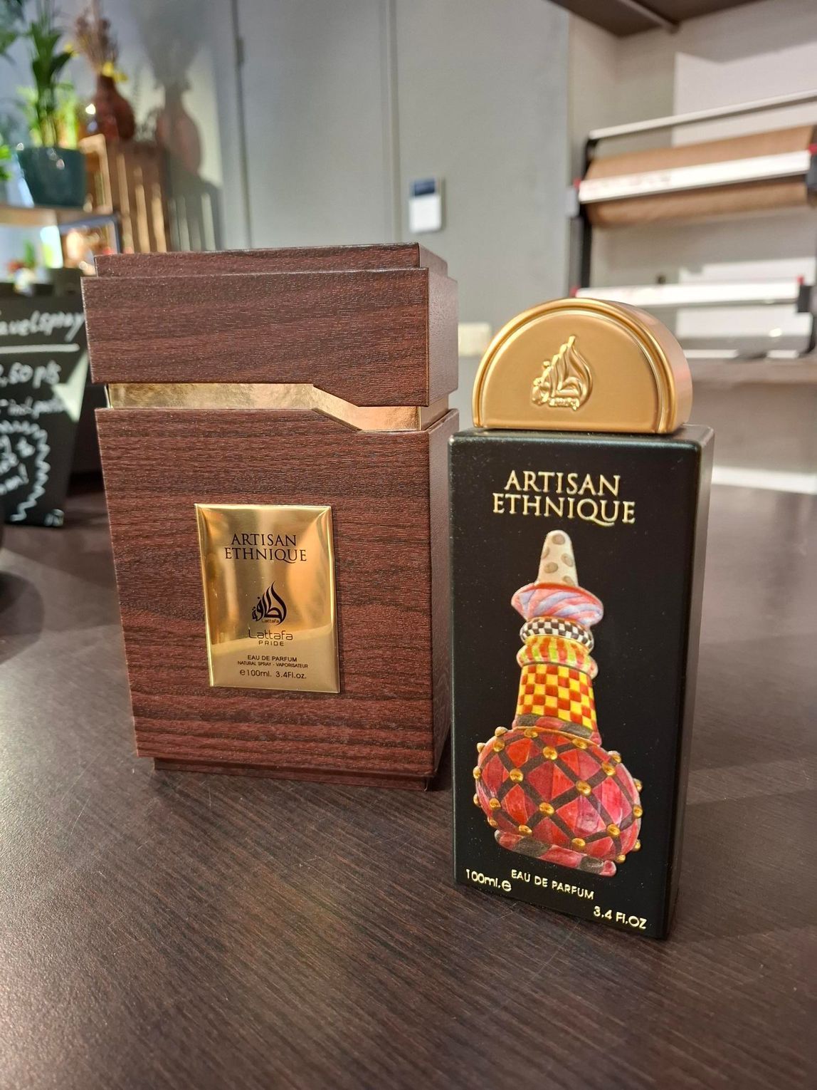 Een fles parfum van Artisan Ethnique met de bijbehorende houten doos op een tafel, met een groene plant op de achtergrond.