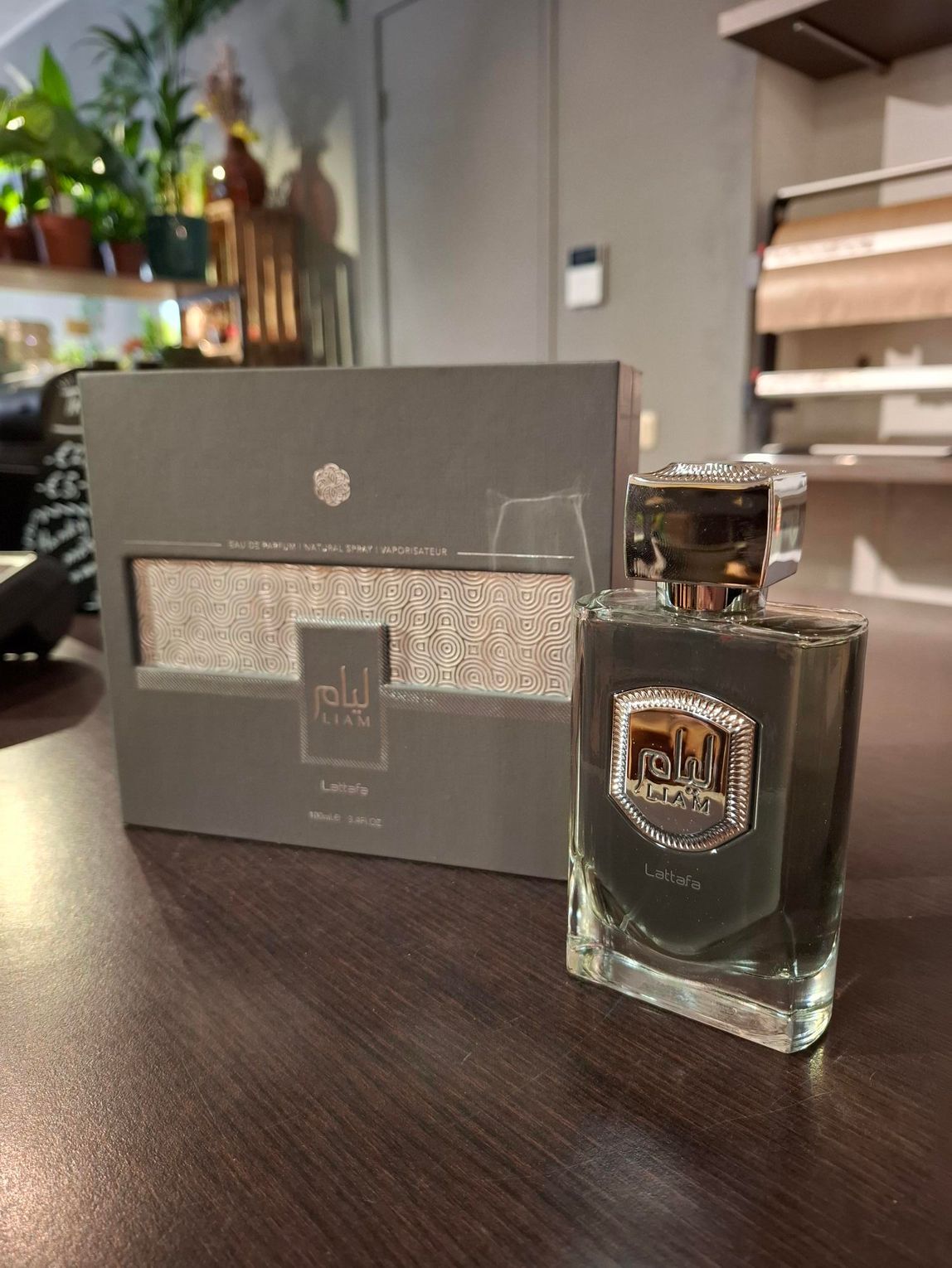 Een flesje donkergekleurde parfum met een metallic etiket staat op een houten ondergrond naast de bijpassende grijze doos.