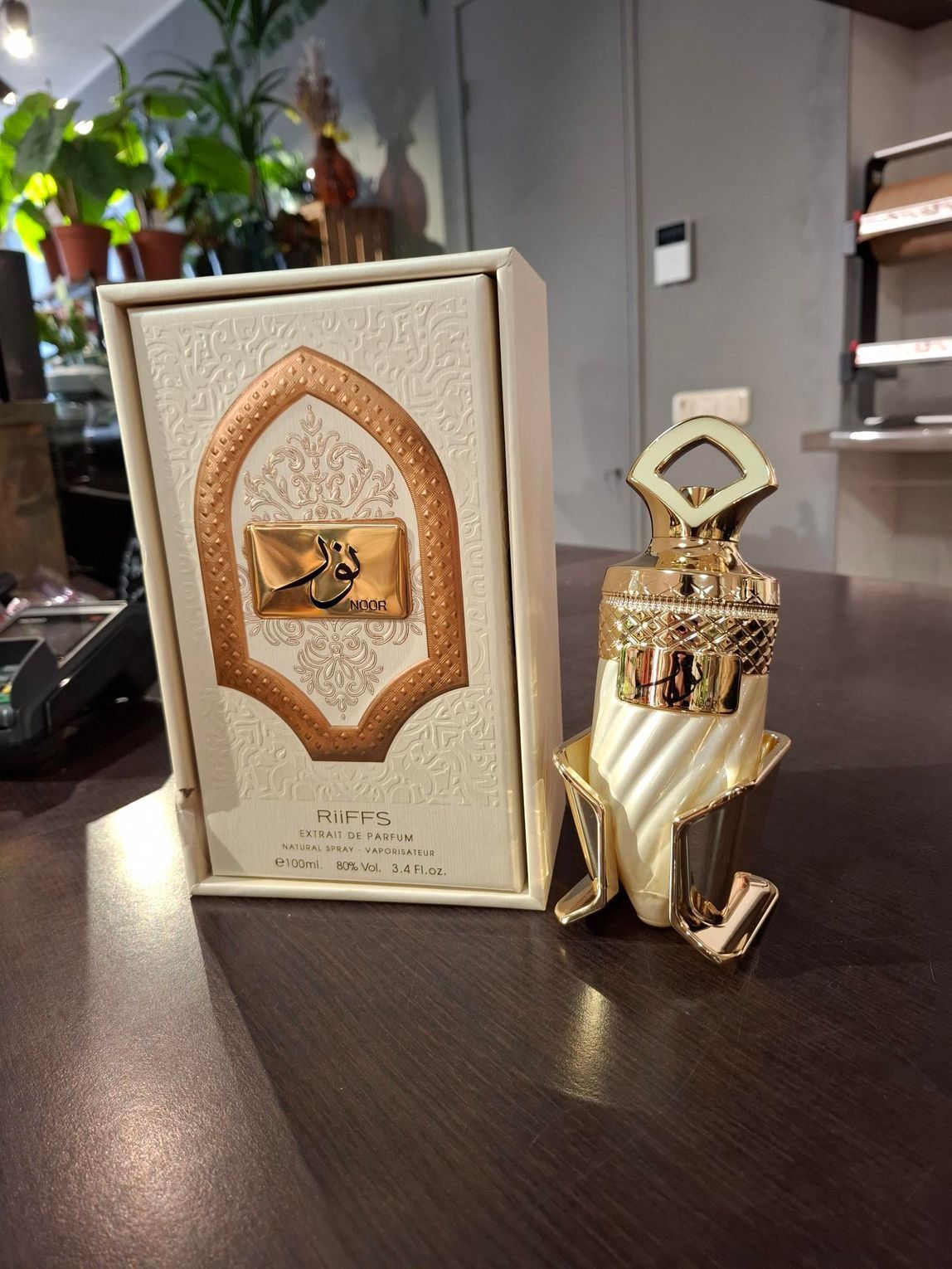 Een sierlijke parfumfles in wit en goud staat naast de bijpassende decoratieve geschenkverpakking op een donkere ondergrond.