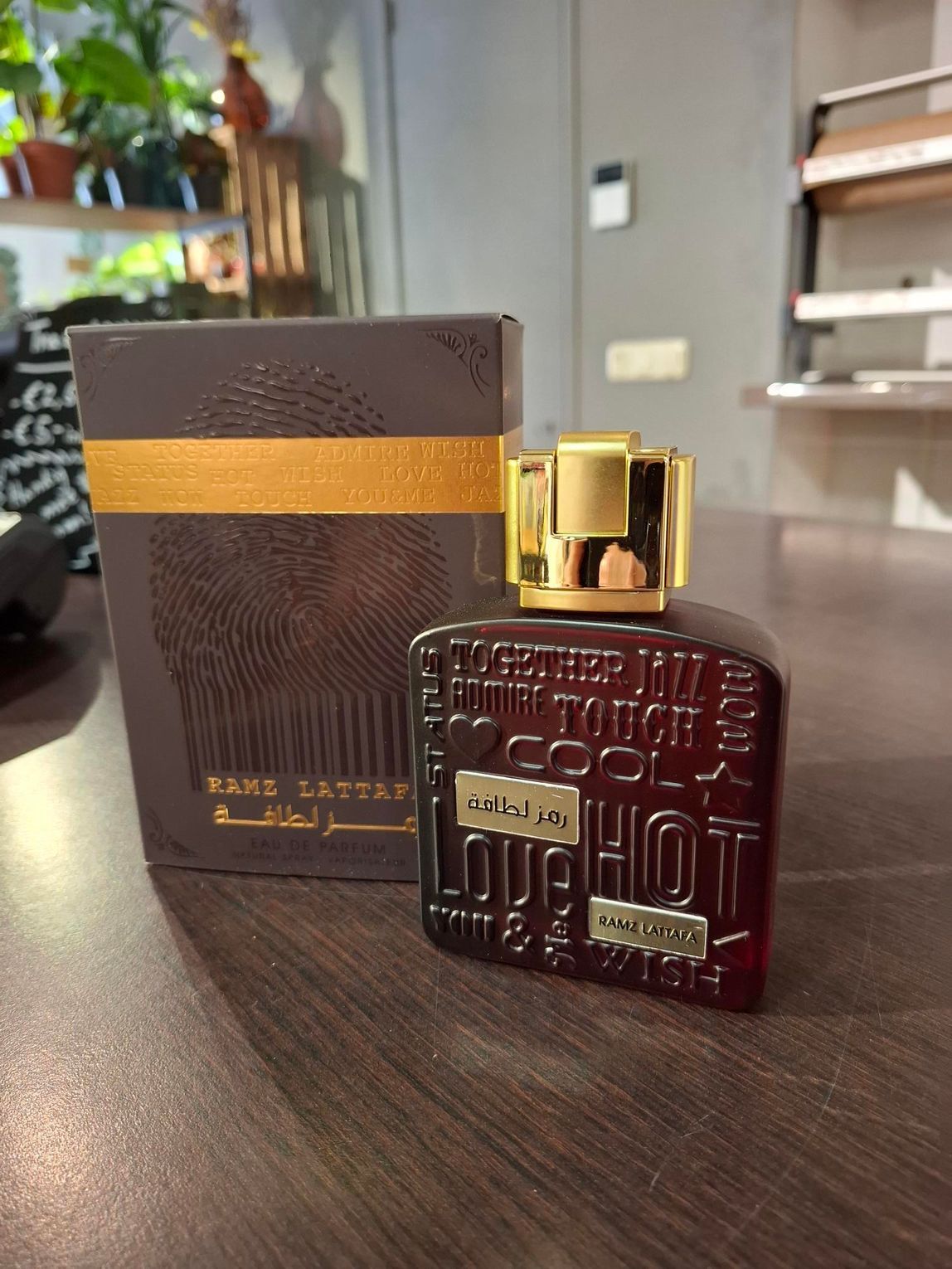 Een donkerrode, rechthoekige parfumfles met reliëftekst staat naast de bijpassende bruine doos op een donkerhouten ondergrond.