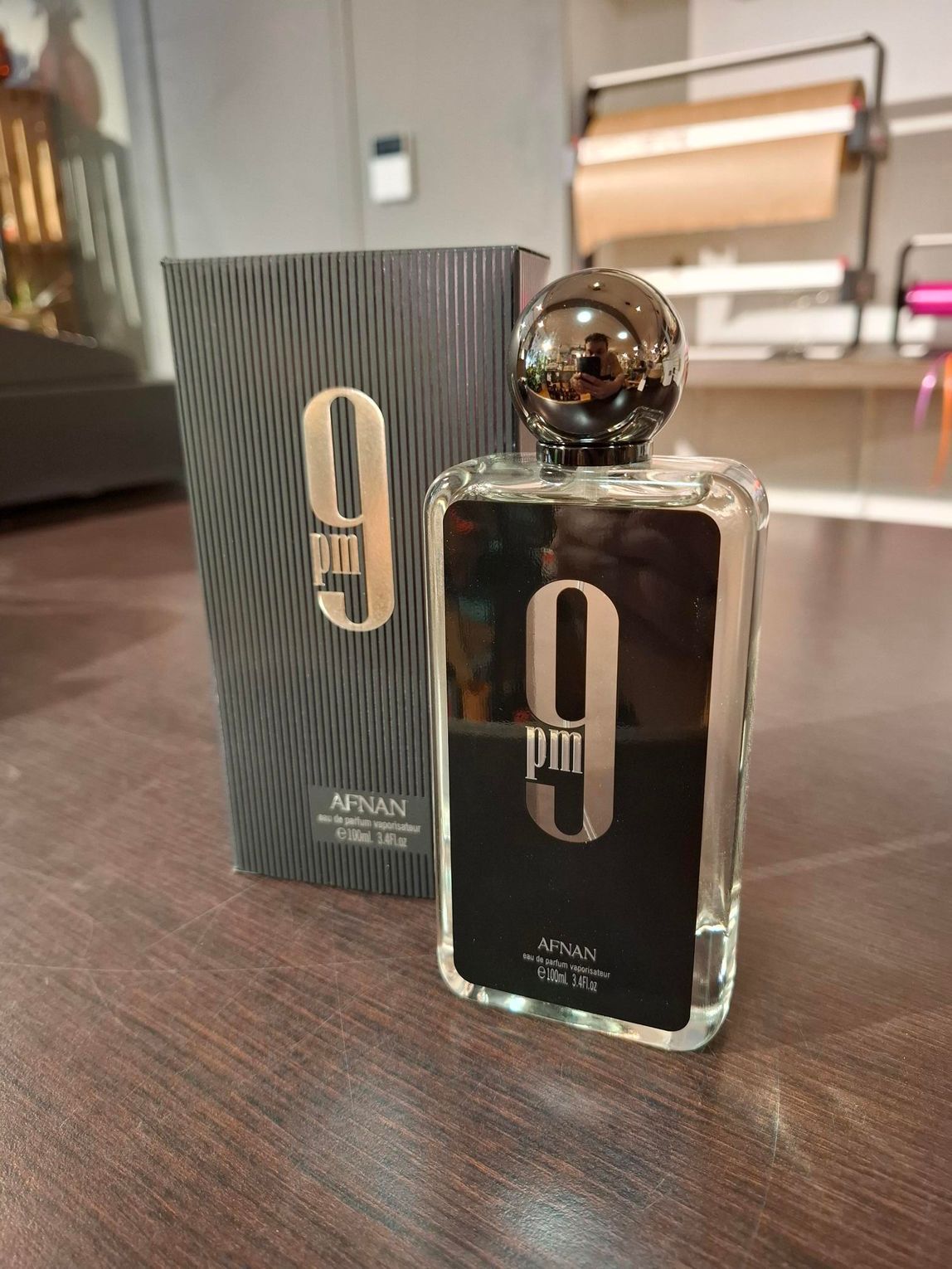 Een fles Afnan 9 PM eau de cologne en de bijbehorende zwarte doos met patroon staan ​​op een houten ondergrond.