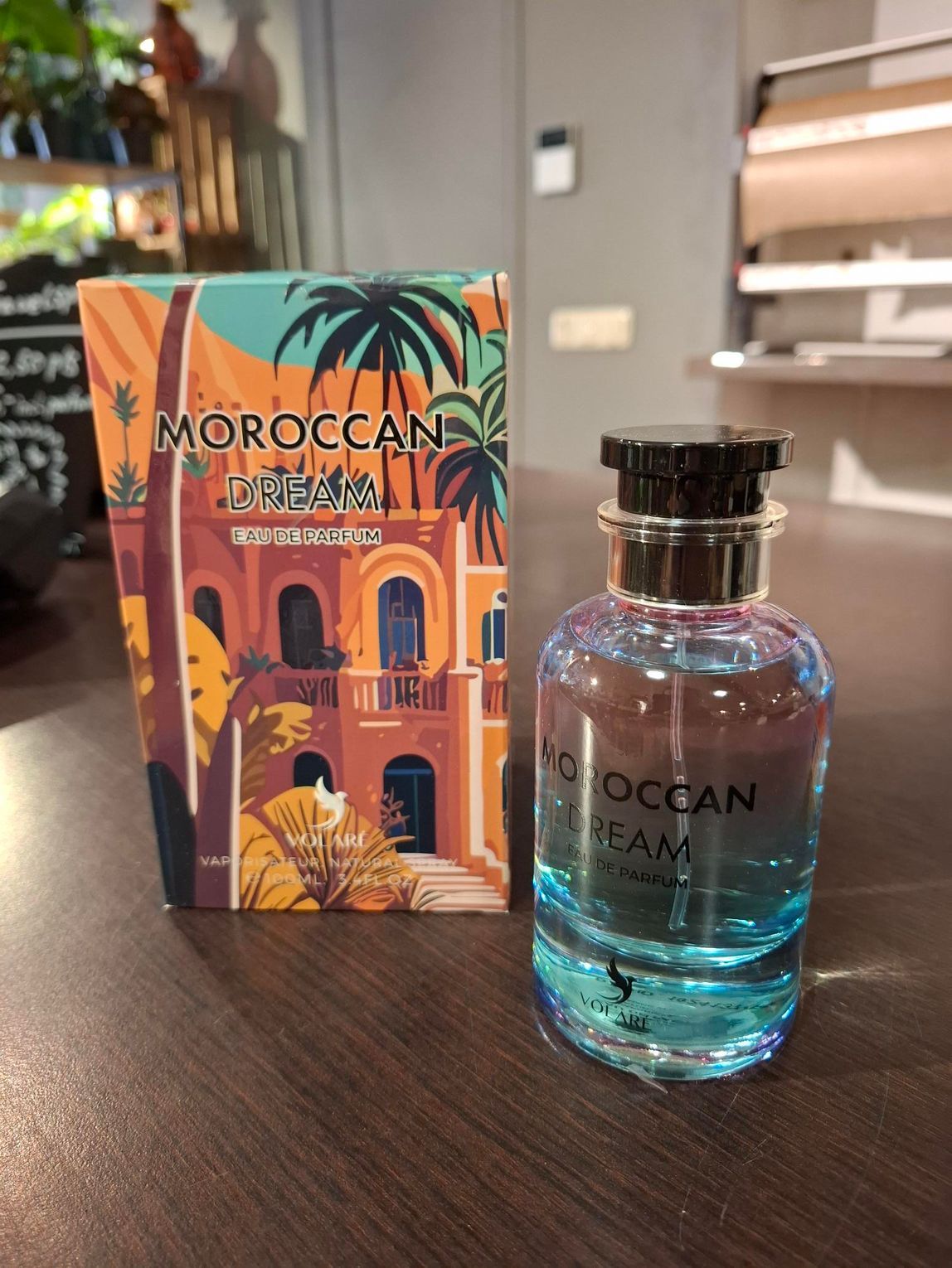 Een flesje Moroccan Dream-parfum met blauwe vloeistof staat naast de geïllustreerde, warmgetinte doos op een houten tafel.