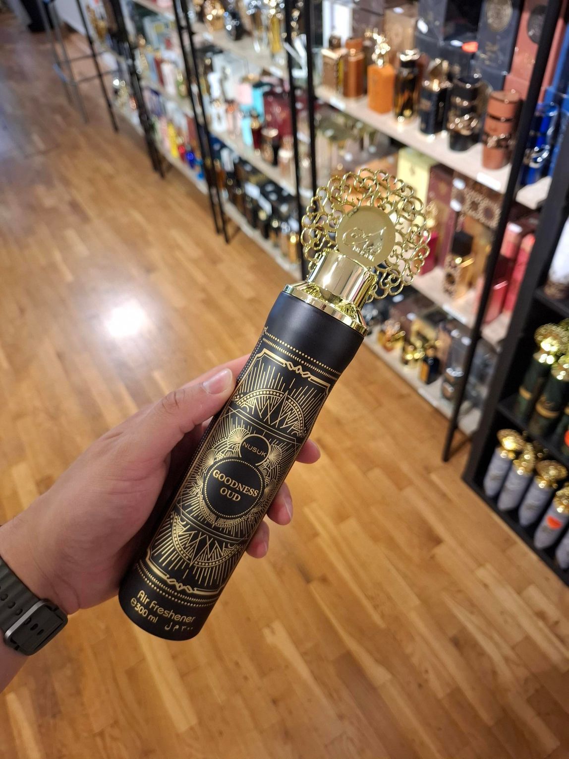 In een winkelgang met schappen vol geurproducten houdt een hand een goud-zwart decoratief parfumflesje vast.