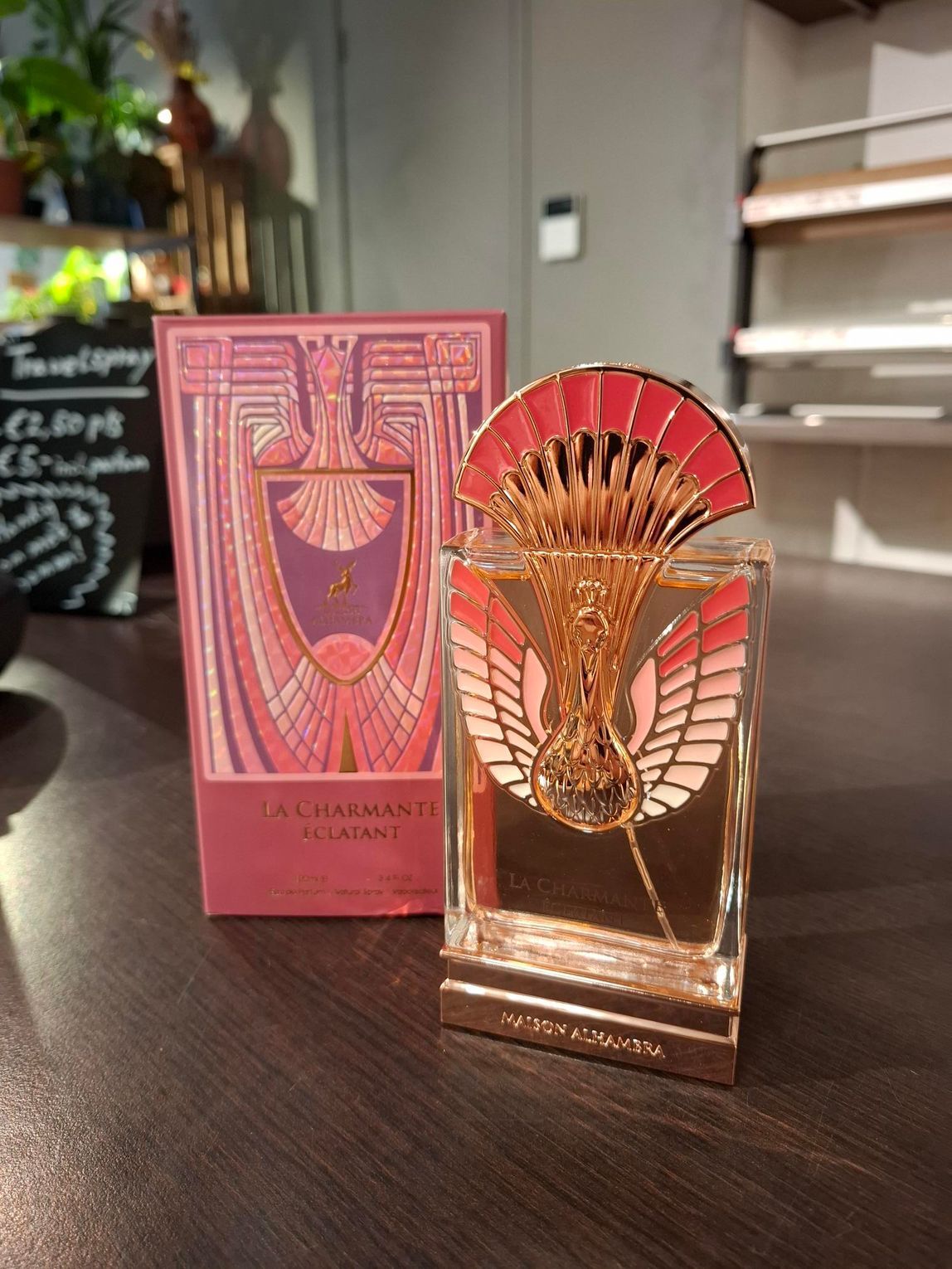 Een rechthoekig parfumflesje met een roze dop met vleugels staat naast de bijpassende roze doos in art-decostijl op een tafel.