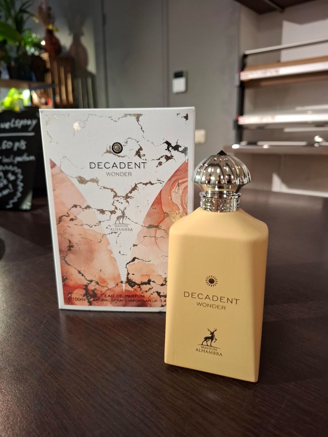 Een flesje parfum Decadent Noir naast de bijpassende doos met marmerpatroon, op een donkerhouten ondergrond.