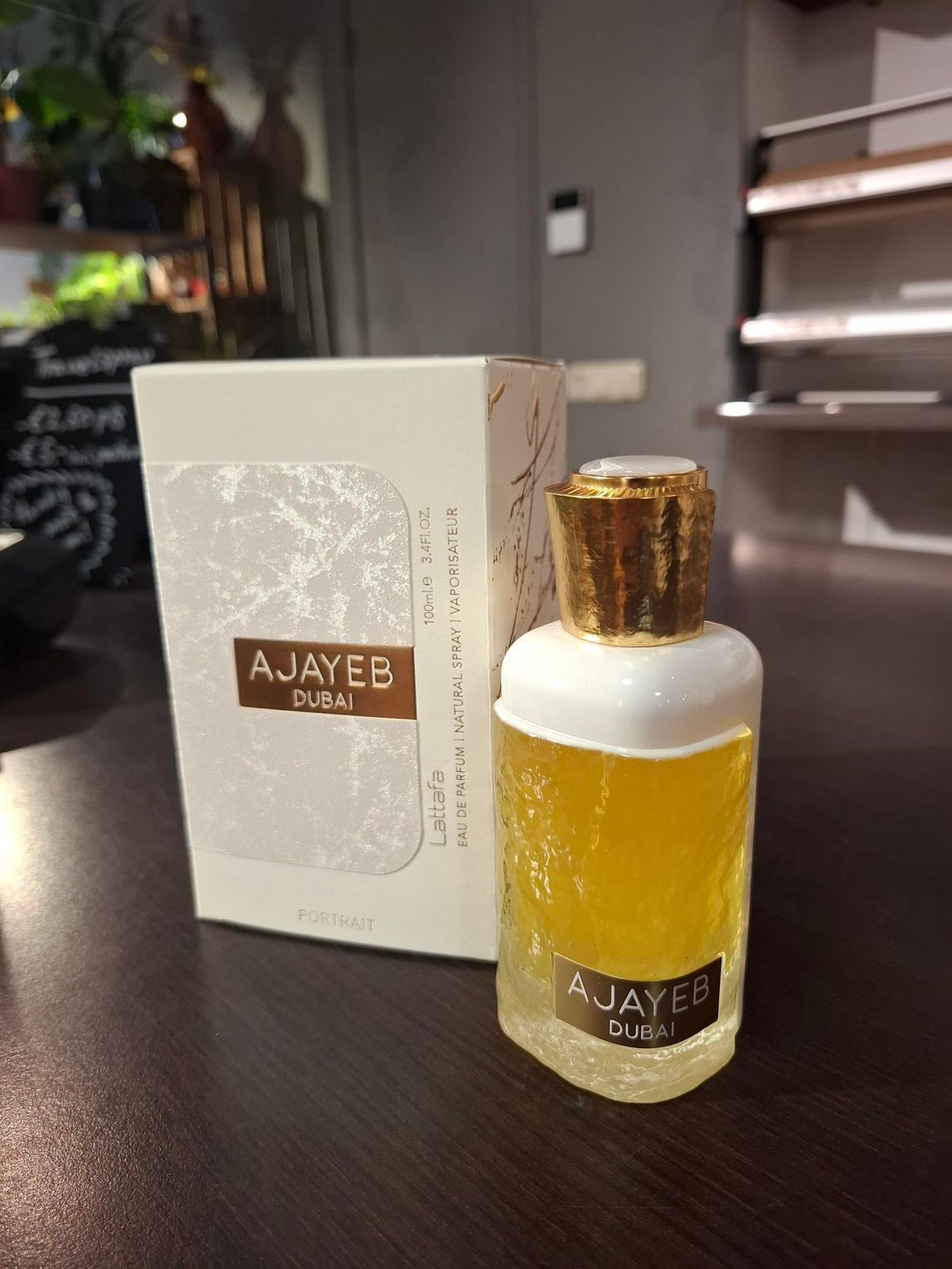 Een fles Ajayeb Dubai-parfum met de bijbehorende witte en gouden doos staat op een donkere ondergrond in een kamer.