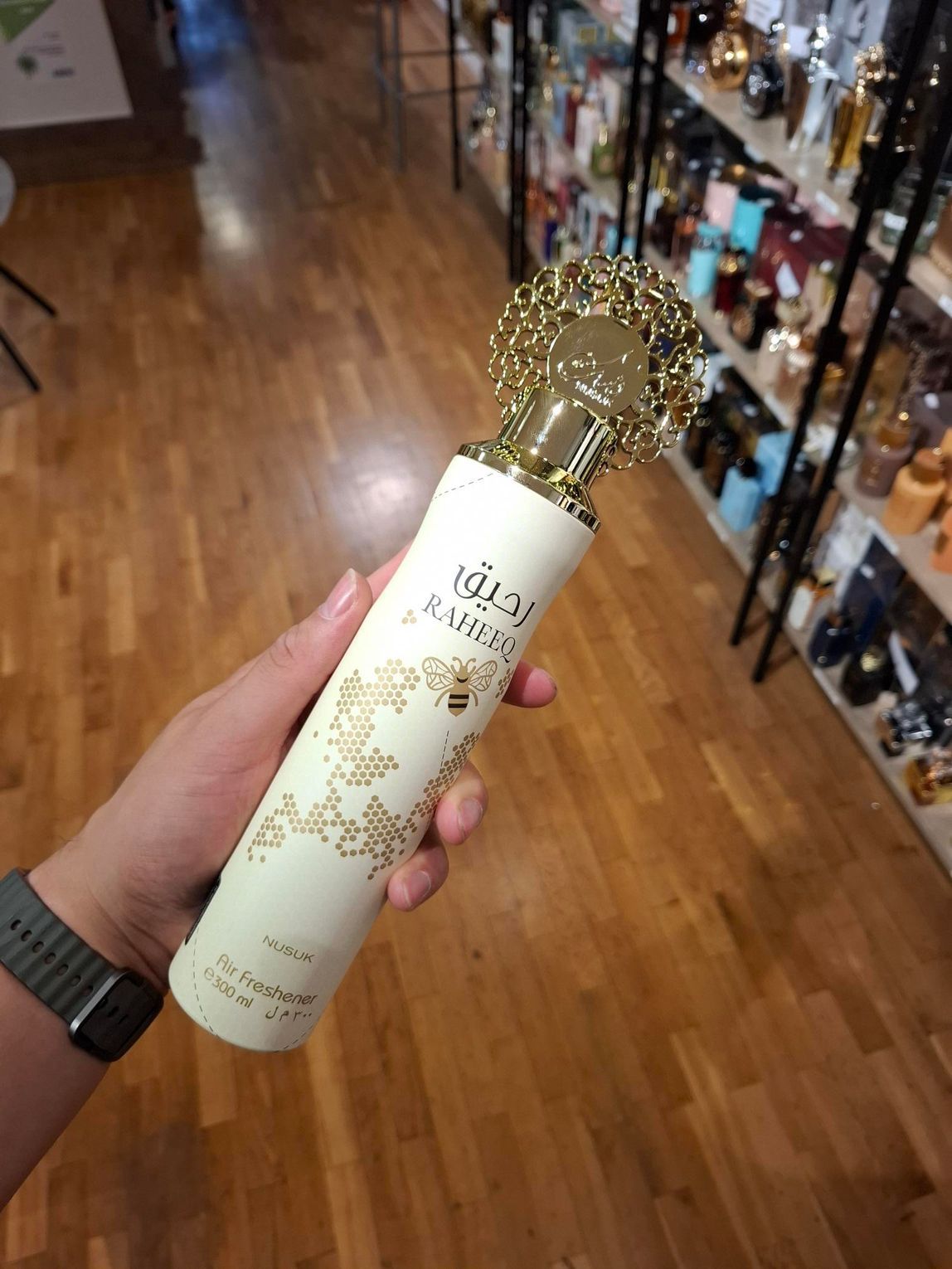 In een winkelgang houdt een hand een crèmekleurig parfumflesje van Rasasi vast, met een decoratieve gouden dop en bloemmotieven.