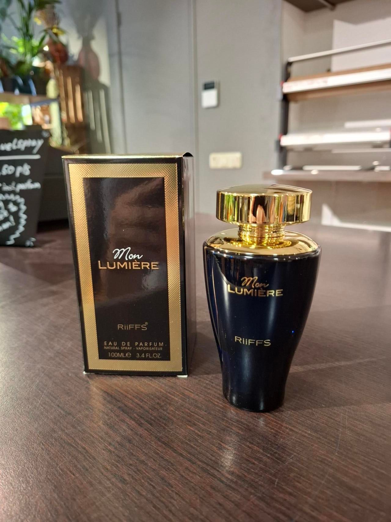 Een donkerblauwe fles Mon Lumière-parfum met een gouden dop staat naast de bijpassende goud-zwarte doos op een houten tafel.