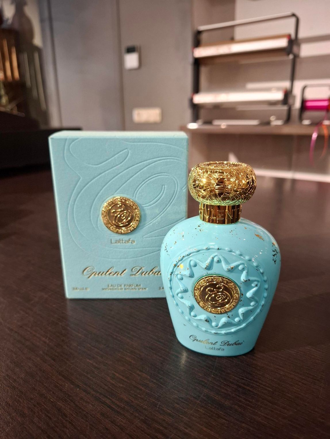 Een lichtblauwe parfumfles en de bijbehorende doos met gedetailleerde, gestructureerde gouden emblemen op een donkerhouten ondergrond.