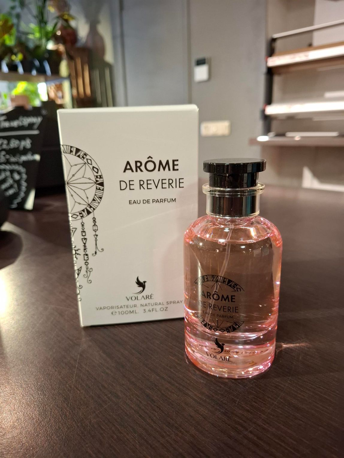 Een flesje roze parfum Arôme de Reverie en de bijbehorende witte verpakking staan ​​op een donkerhouten oppervlak in een winkel.