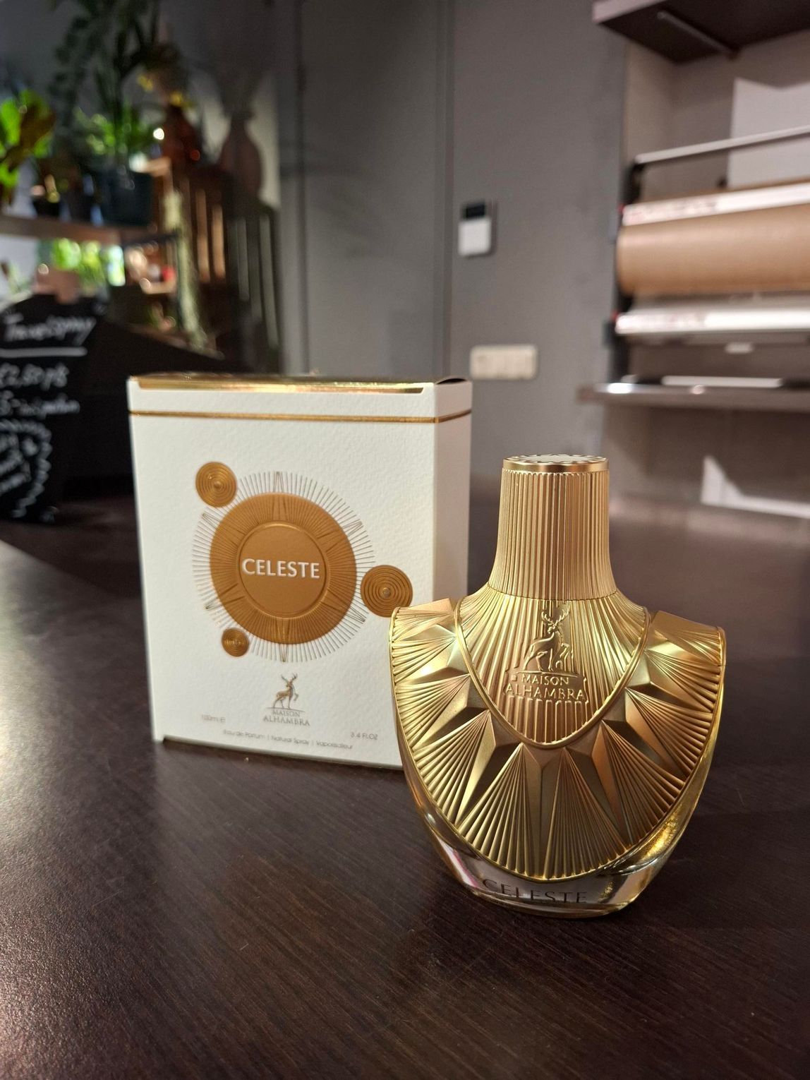 Een goudkleurige, fraai vormgegeven parfumfles staat naast de wit-gouden doos op een donkere ondergrond in een schemerig verlichte kamer.