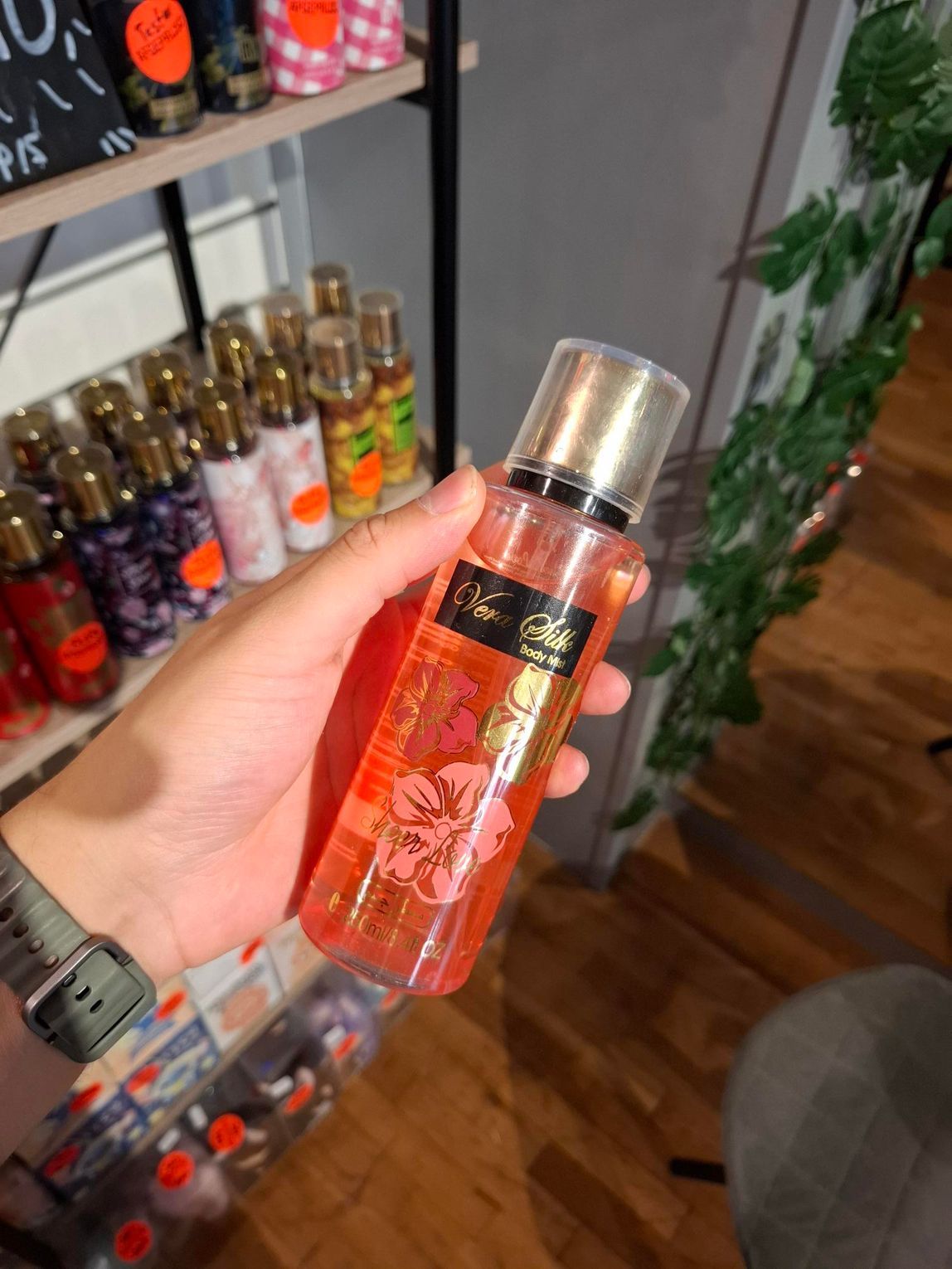 Een hand houdt een koraalkleurige Victoria's Secret body mist sprayflacon vast met bloemenmotief, in een winkelomgeving.