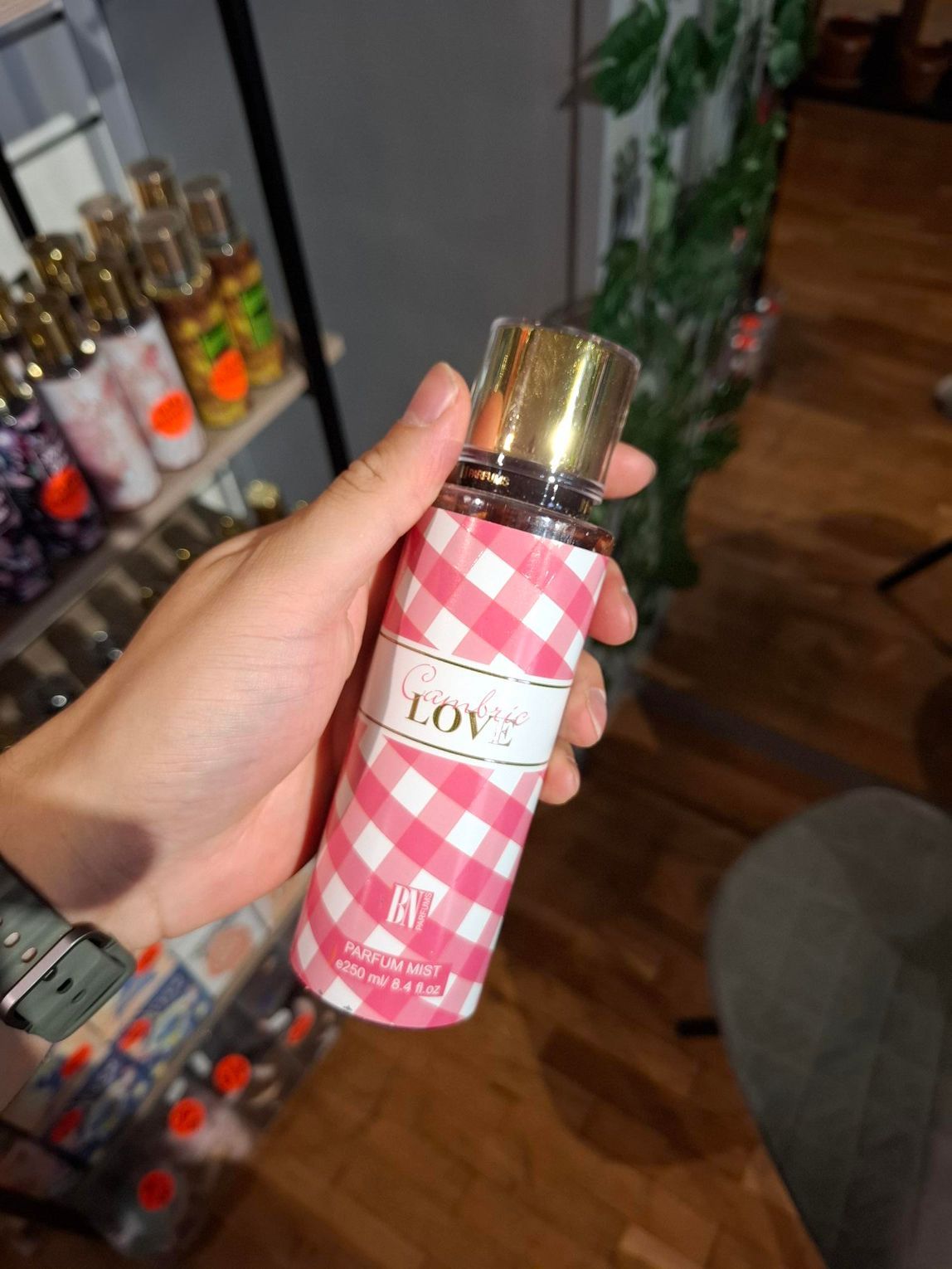In een winkelgang houdt een hand een flesje body mist vast met een roze en witte ruitjesprint en een gouden dop.