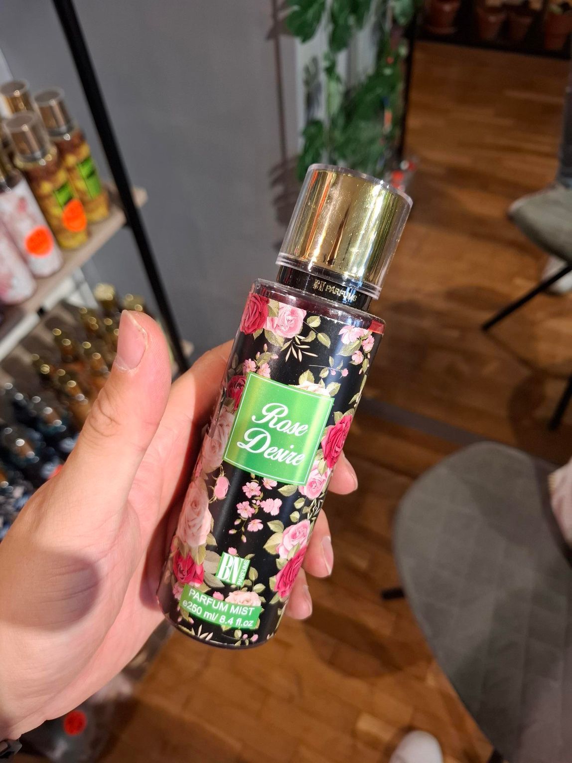 In een winkel houdt een hand een flesje body mist met bloemenprint vast, waarop 