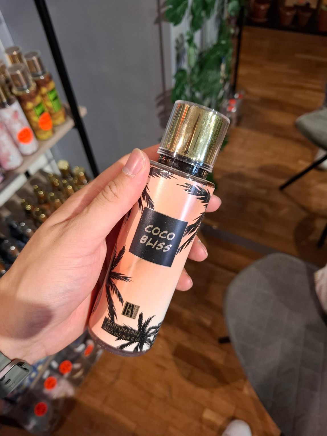 In een winkel houdt een hand een perzikkleurige spuitfles vast met een gouden dop en een palmboommotief, waarop 