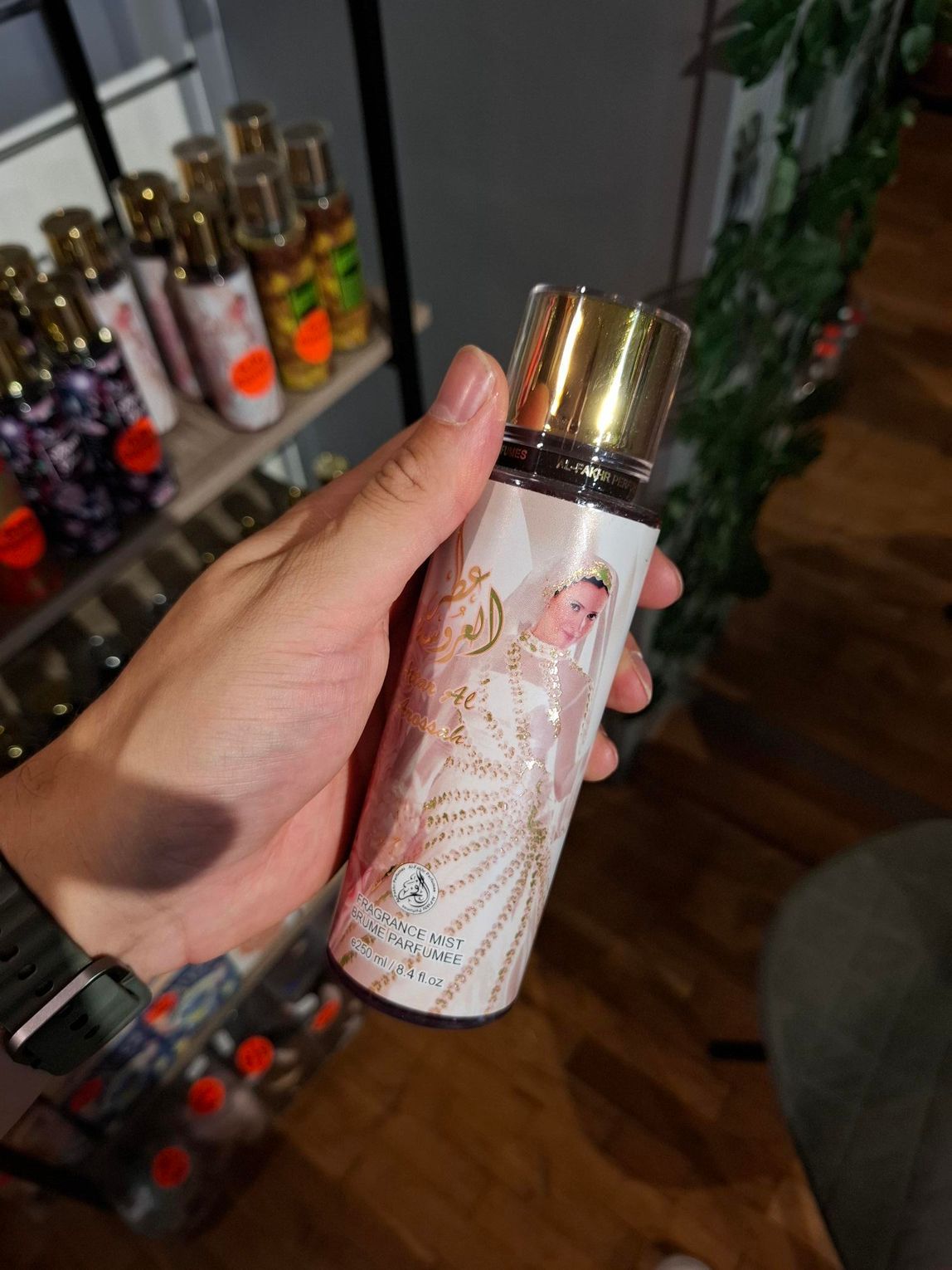 Een hand houdt een parfumflesje vast met een gouden dop en een etiket waarop een afbeelding staat van een figuur in een met kralen versierde jurk.