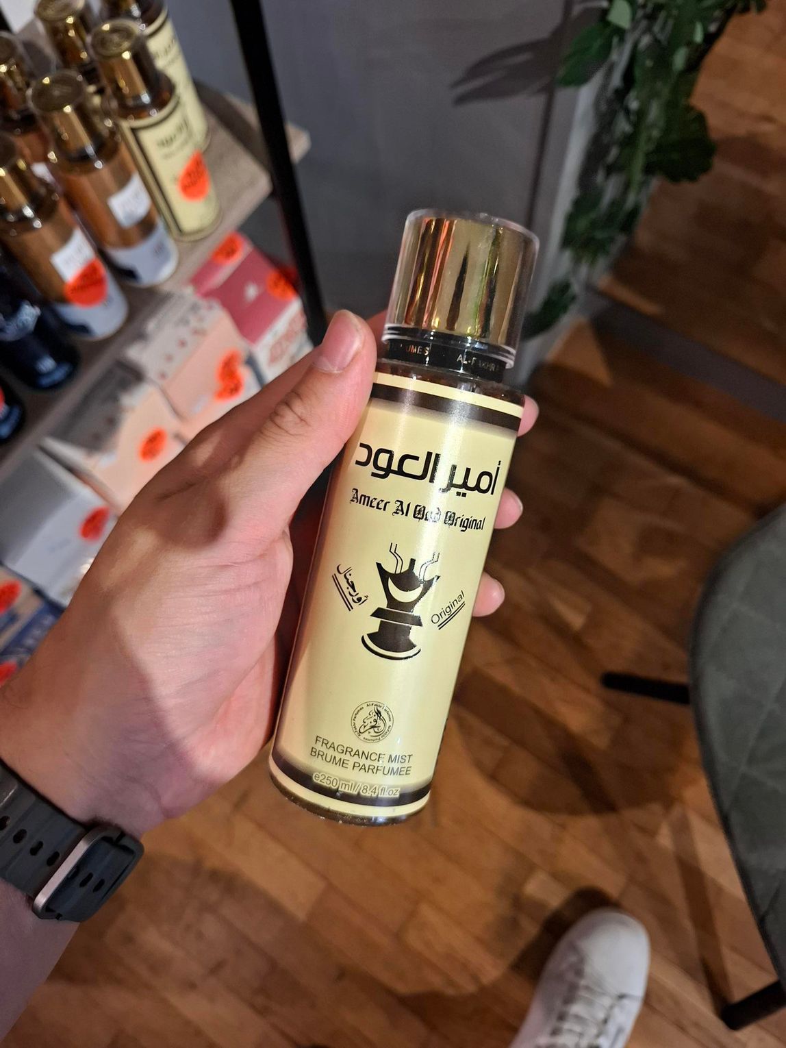 In een winkel houdt een hand een flesje Amber Oud-parfum vast, op de achtergrond zijn andere parfumflesjes te zien in de schappen.