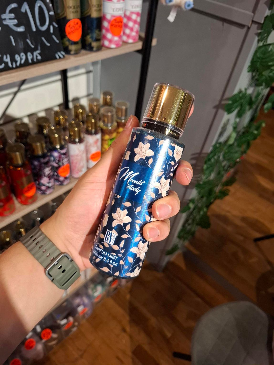 In een winkel, met op de achtergrond schappen vol parfumflesjes, houdt een hand een donkerblauwe fles body mist met bloemenprint vast.