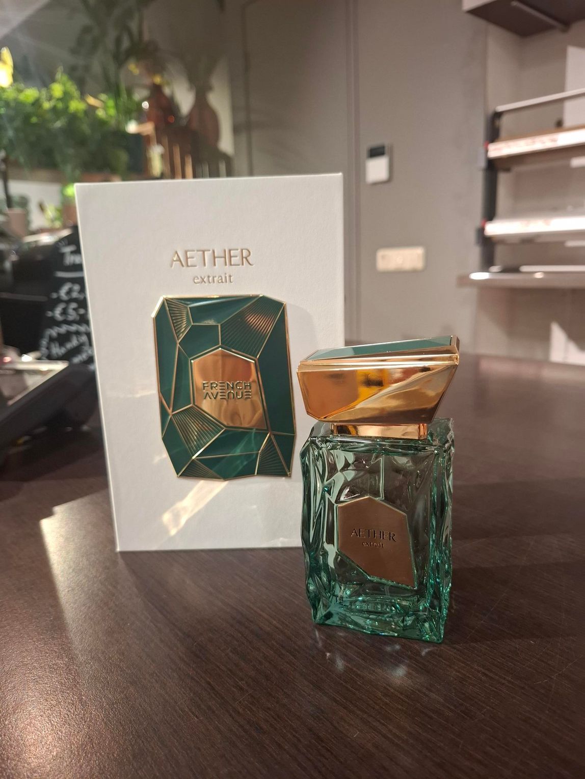 Een wit parfumdoosje met een groen geometrisch embleem staat naast een glazen flesje Aether-parfum op een donkere tafel.