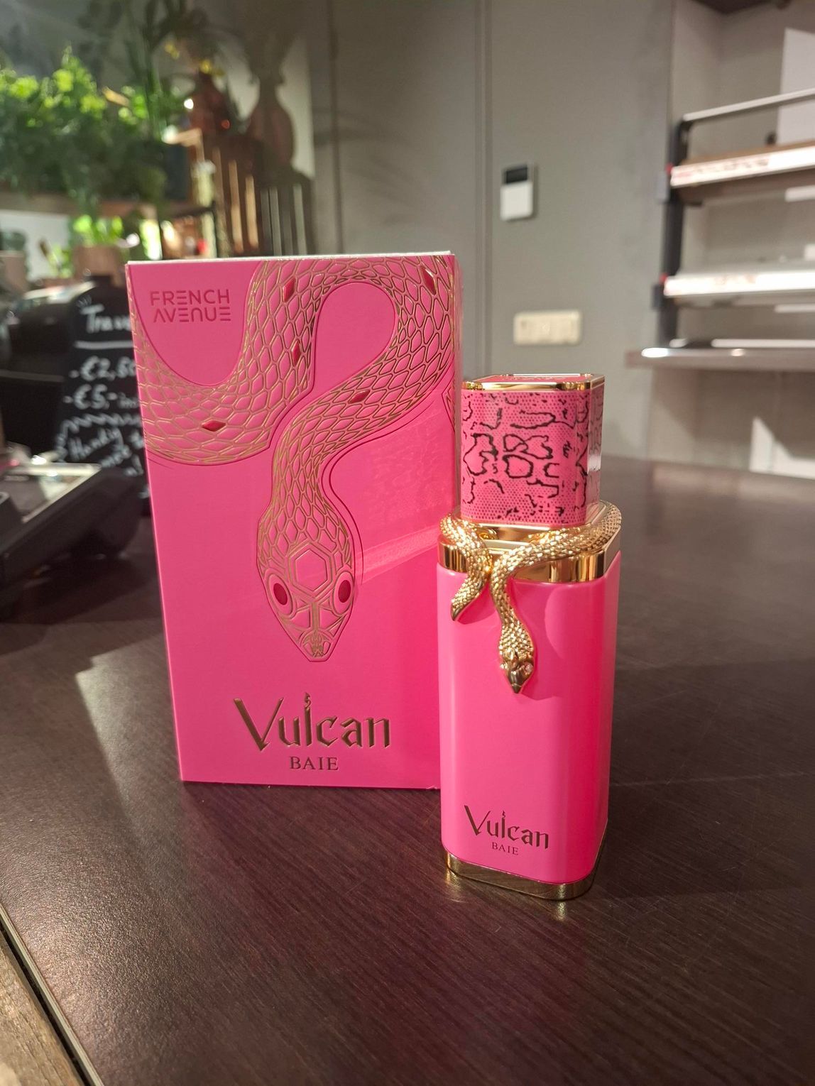 Een felroze parfumflesje en het bijbehorende doosje, beide versierd met gouden slangenmotieven, staan ​​op een houten toonbank.