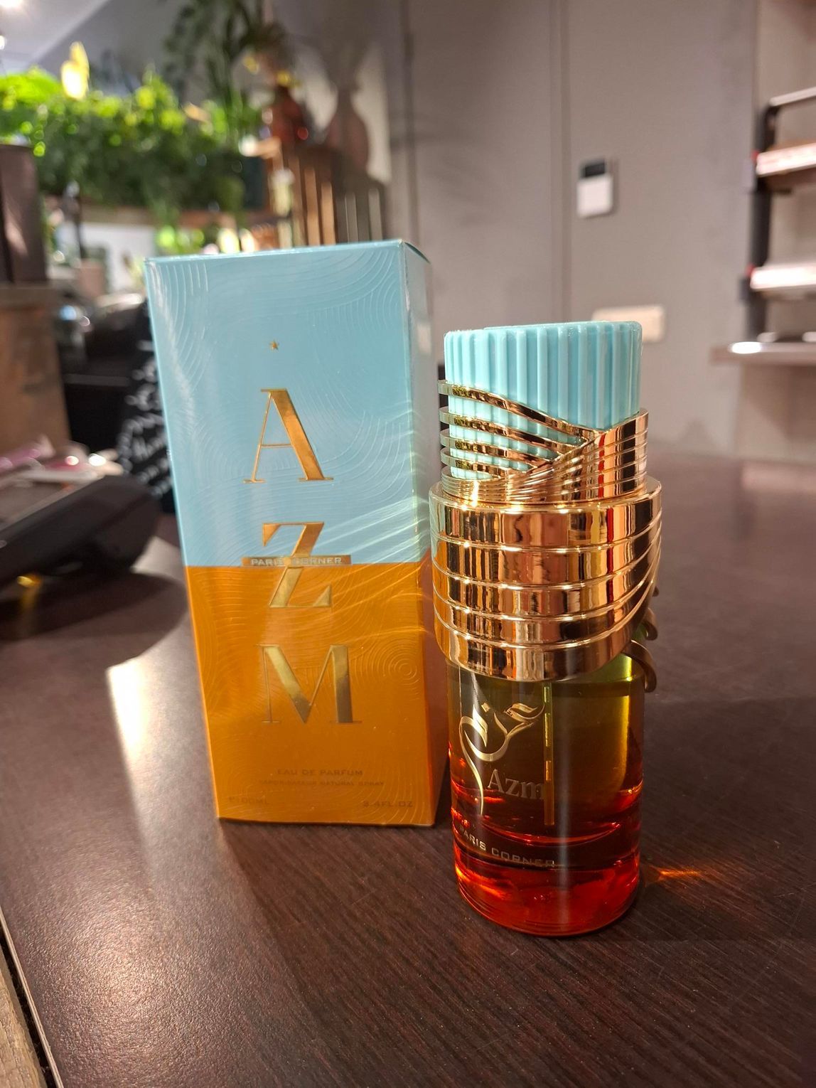 Een flesje Azm-parfum naast de blauw-gele doos, op een houten oppervlak in een winkel.