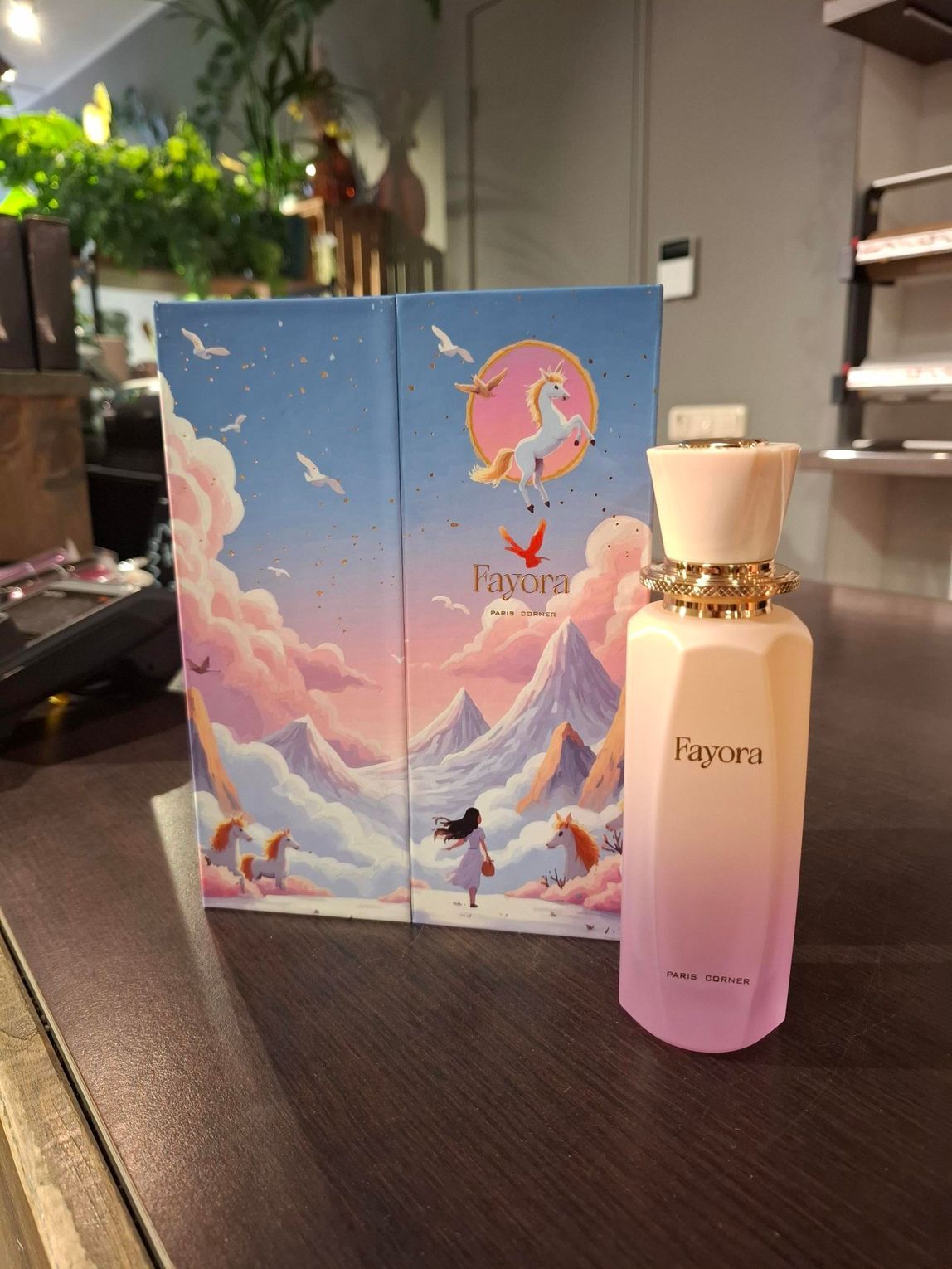 Een parfumflesje met bijbehorend doosje, versierd met een speelse illustratie van een eenhoorn en bergen, staat op een donkerhouten tafel.