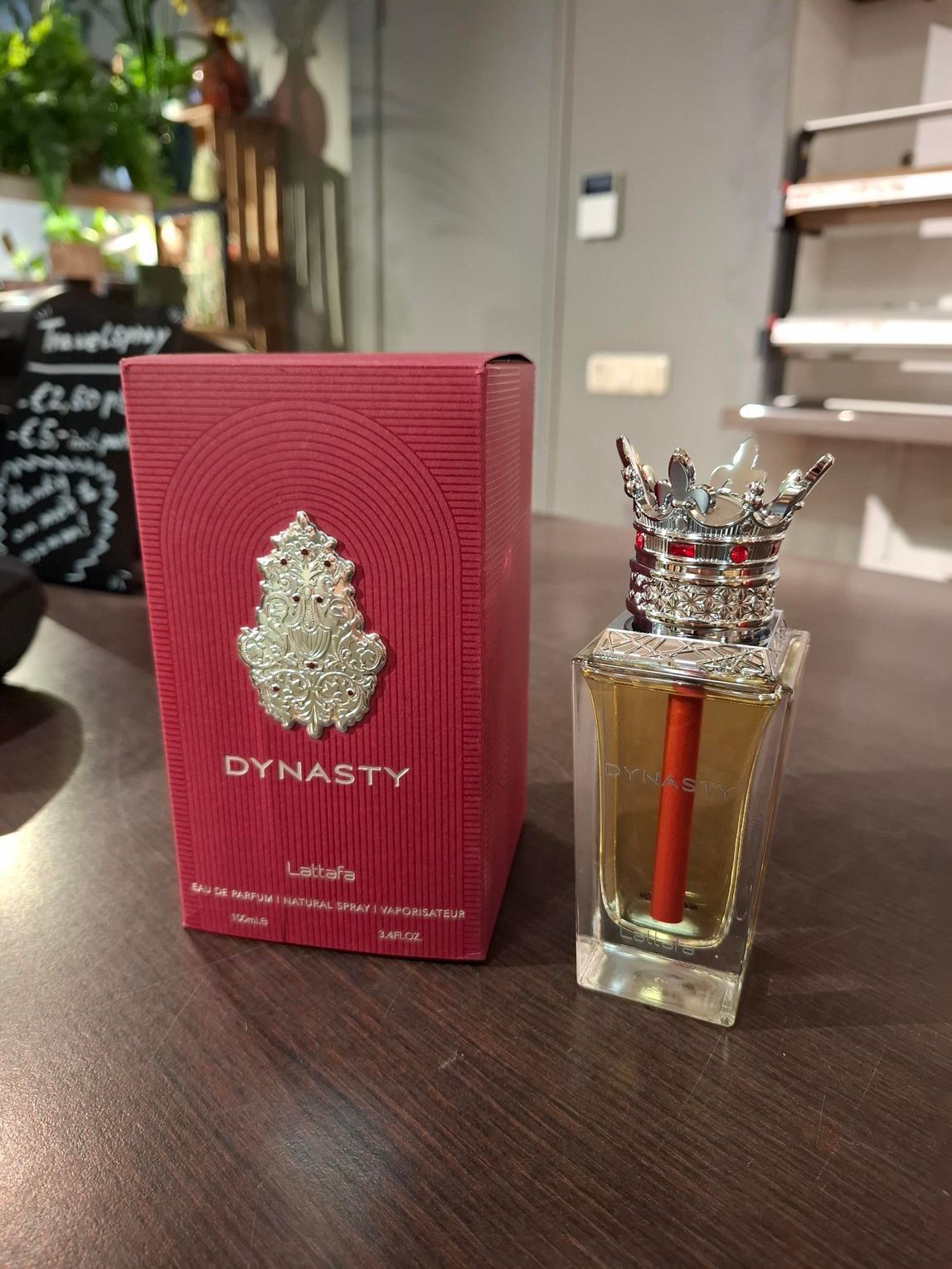Een parfumflesje van Dynasty en de bijbehorende rode, gestructureerde doos met een sierlijk zilveren embleem, staan ​​op een houten ondergrond.