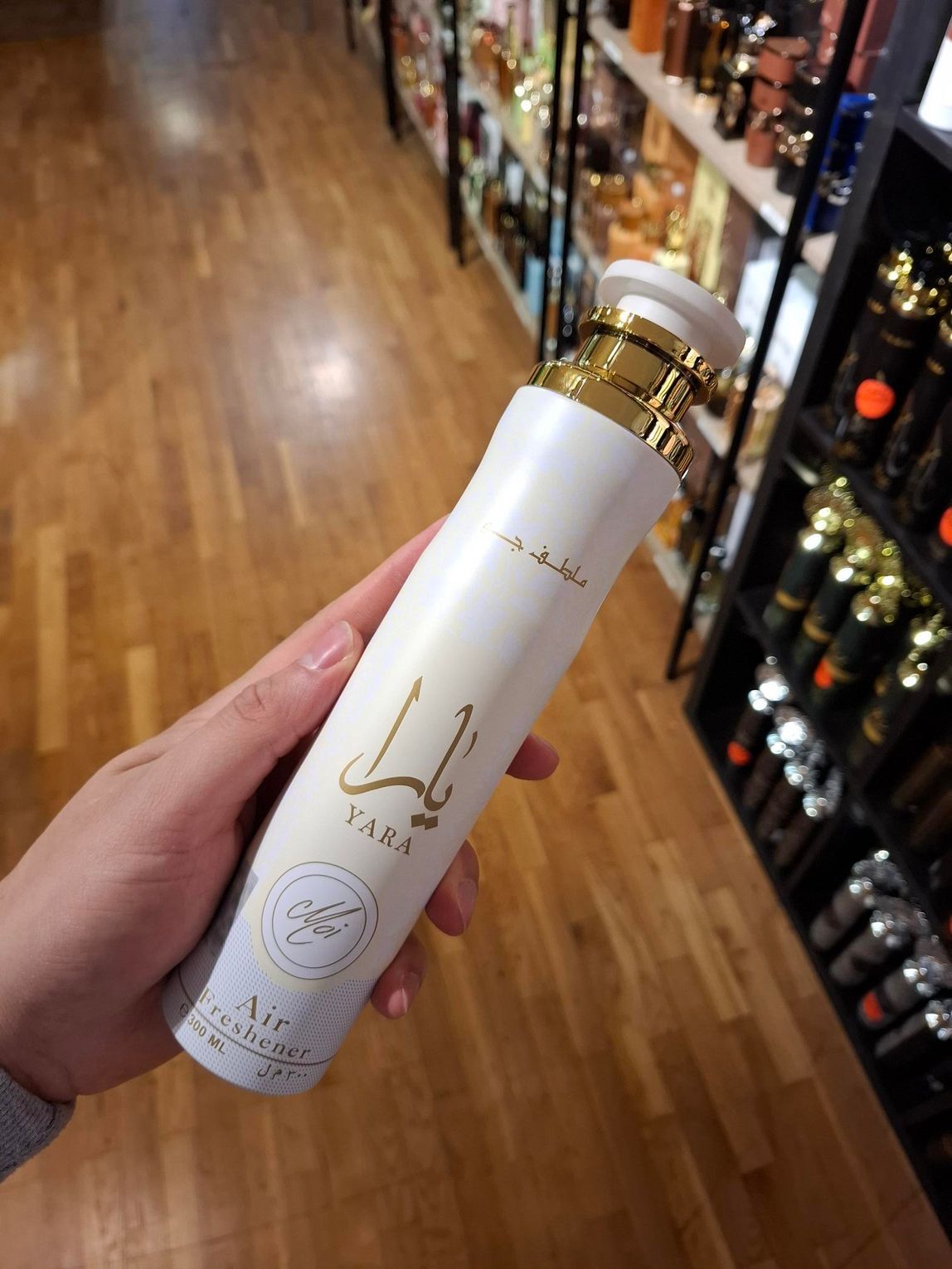 In een winkelgang houdt een hand een hoge, witte geurverstuiver met gouden letters en dop vast.