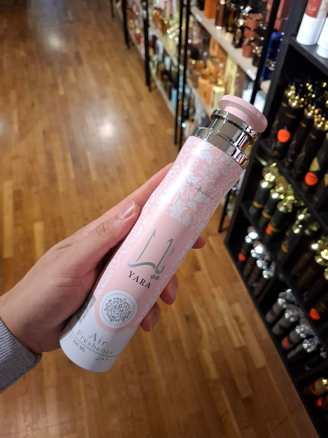 Een hand houdt een lange, cilindrische, lichtroze spuitbus met Lattafa Yara parfum vast, tegen een achtergrond van winkelschappen.