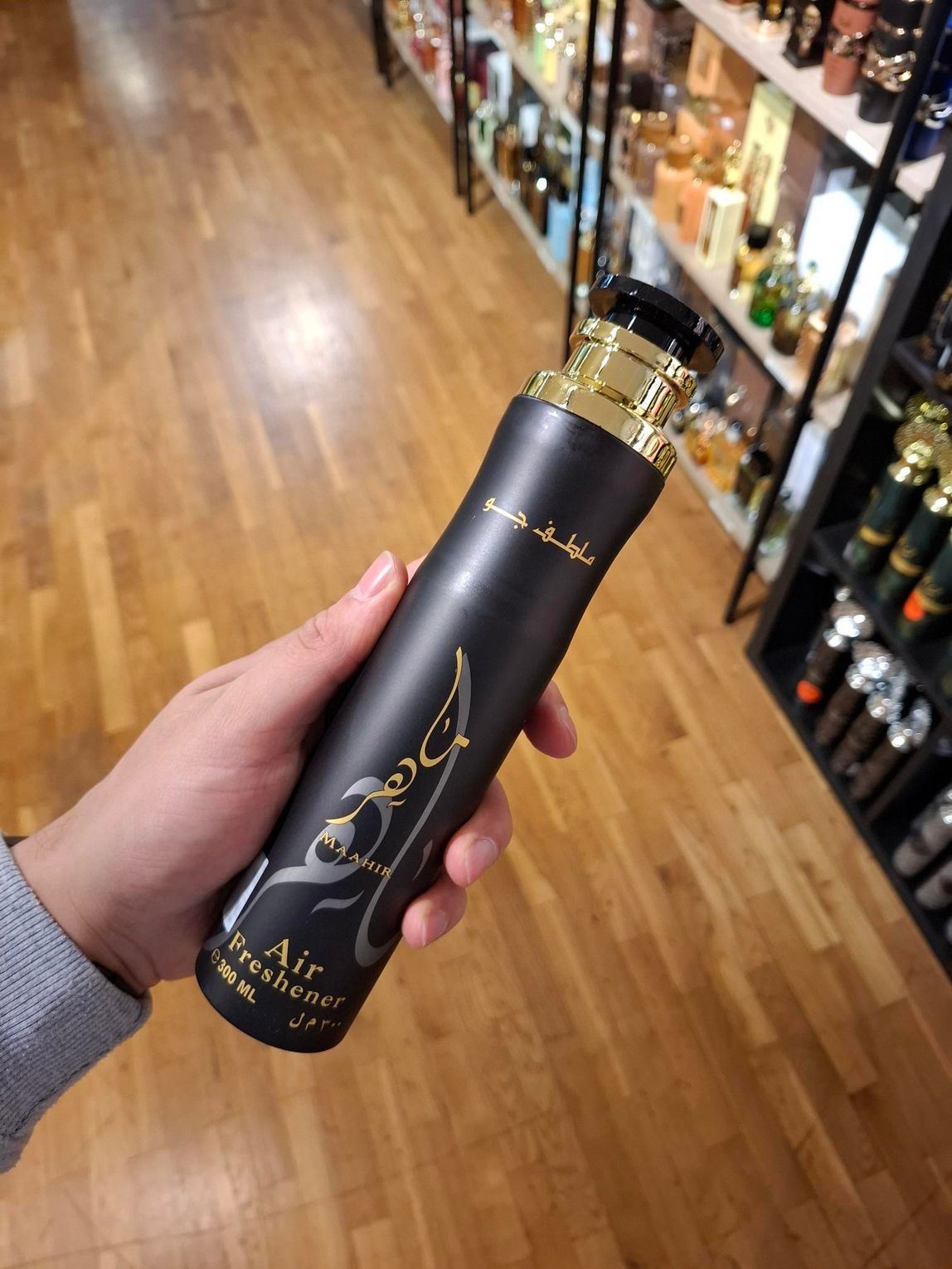 In een winkelgang houdt een hand een slanke, matzwarte luchtverfrisser vast met gouden decoratieve Arabische kalligrafie.
