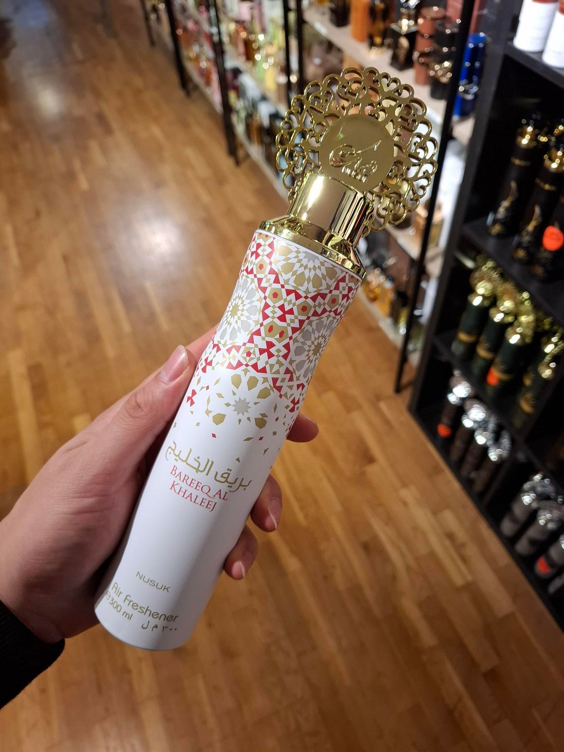 In een winkel houdt een hand een hoge, cilindrische witte parfumfles vast met sierlijke gouden, rode en filigraanpatronen.