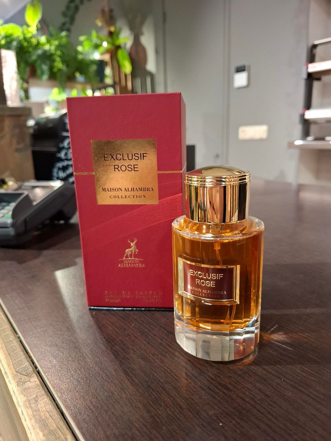 Een fles Exclusive Rose-parfum en de bijbehorende rode doos staan ​​op een donkerhouten ondergrond in een binnenruimte.