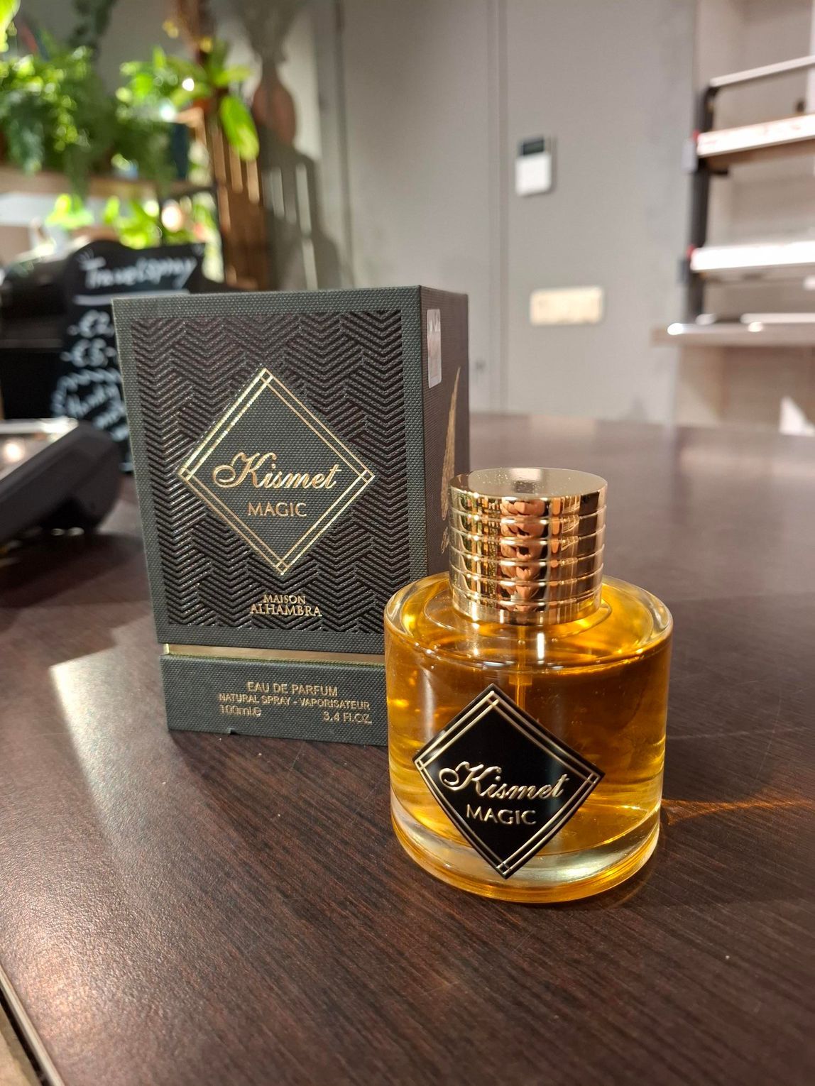 Een flesje Kismet Magic-parfum en de bijbehorende zwarte, gestructureerde doos staan ​​op een donkere houten tafel in een kamer met planten.