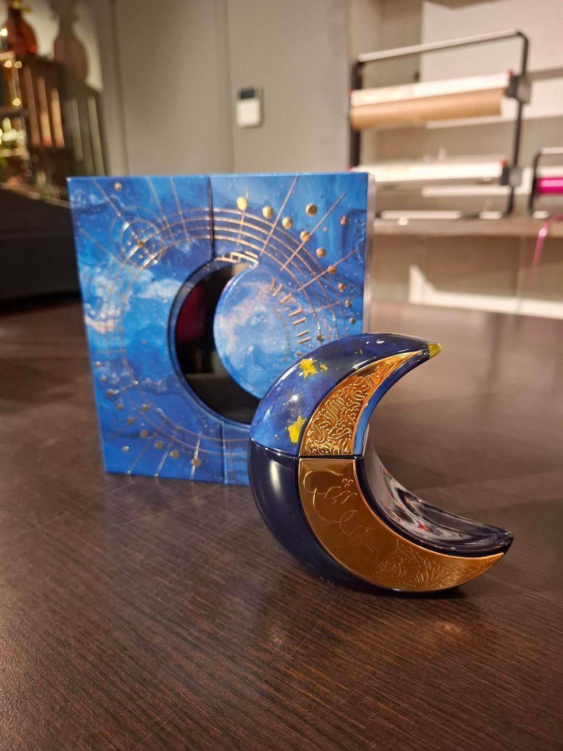 Een parfumflesje in de vorm van een halve maan met gouden glitter, geplaatst voor de bijpassende blauwe doos met hemels thema.